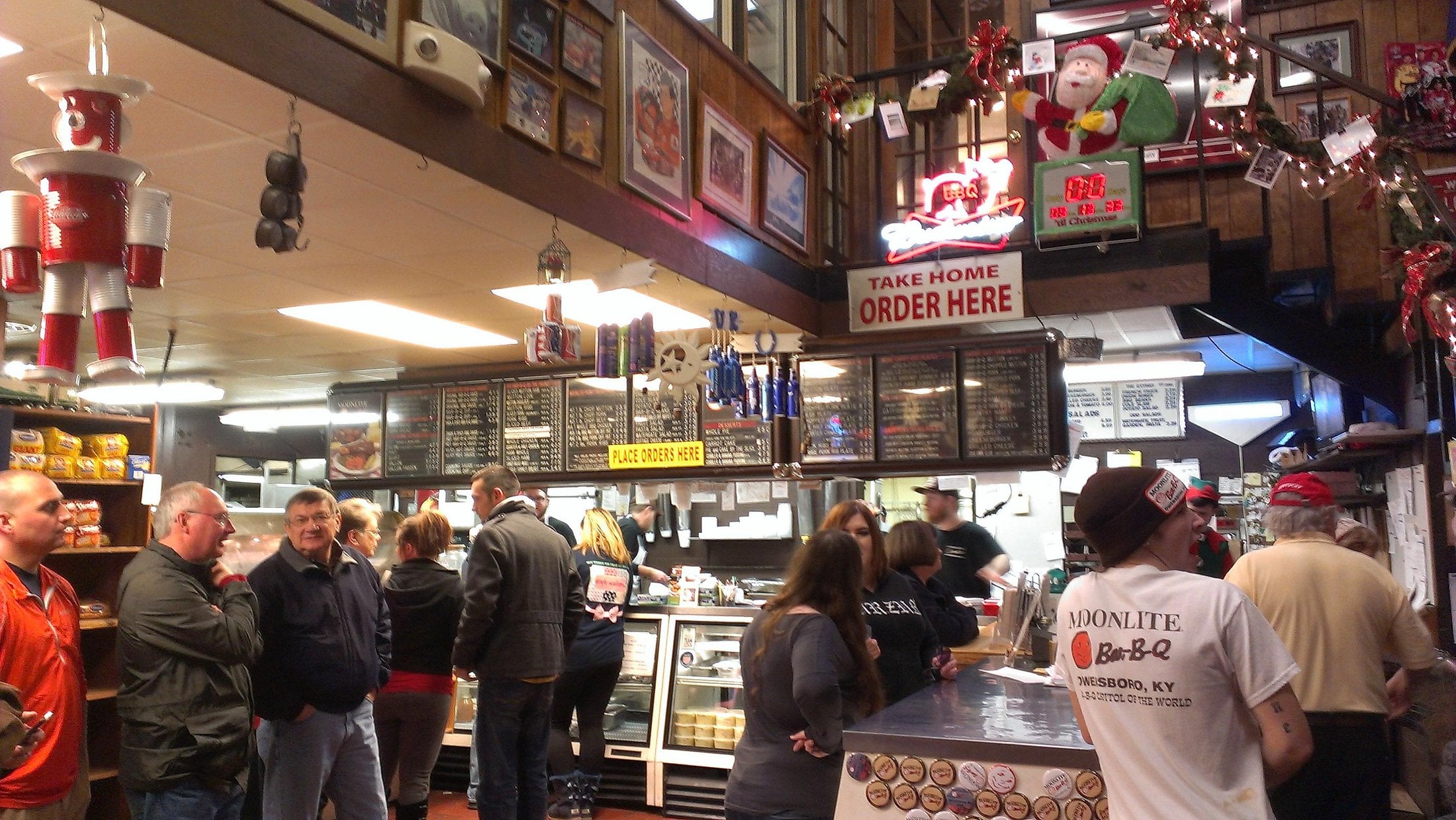 Customers wait in line at a busy barbecue restaurant decorated with festive lights, framed photos, and holiday ornaments. A sign above the counter reads “TAKE HOME ORDER HERE.” Staff work behind the counter.