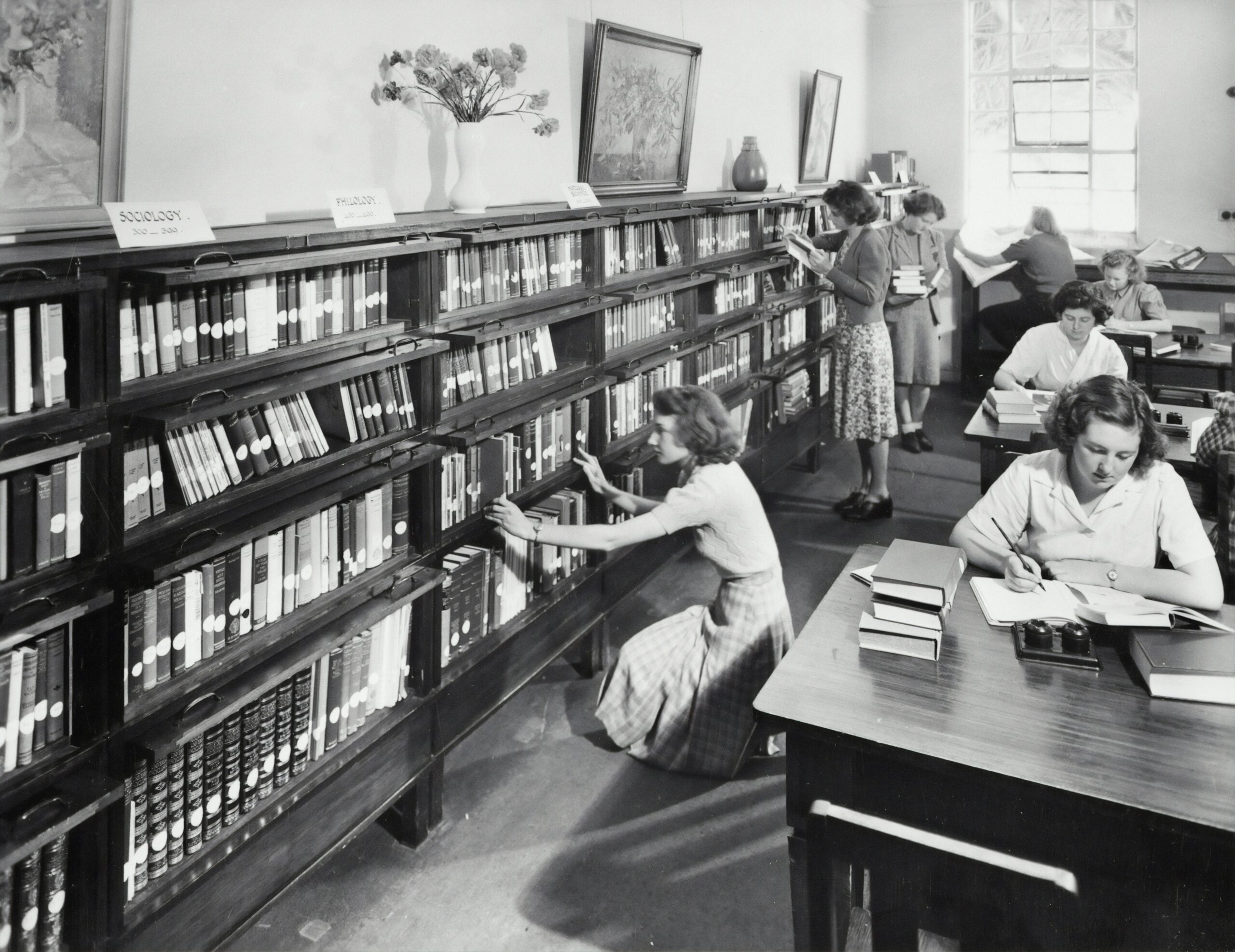 Black-and-white photo of women in a library. Some are shelving or browsing books, while others read or write at tables. Shelves are labeled by subject, and sunlight shines in through a window.