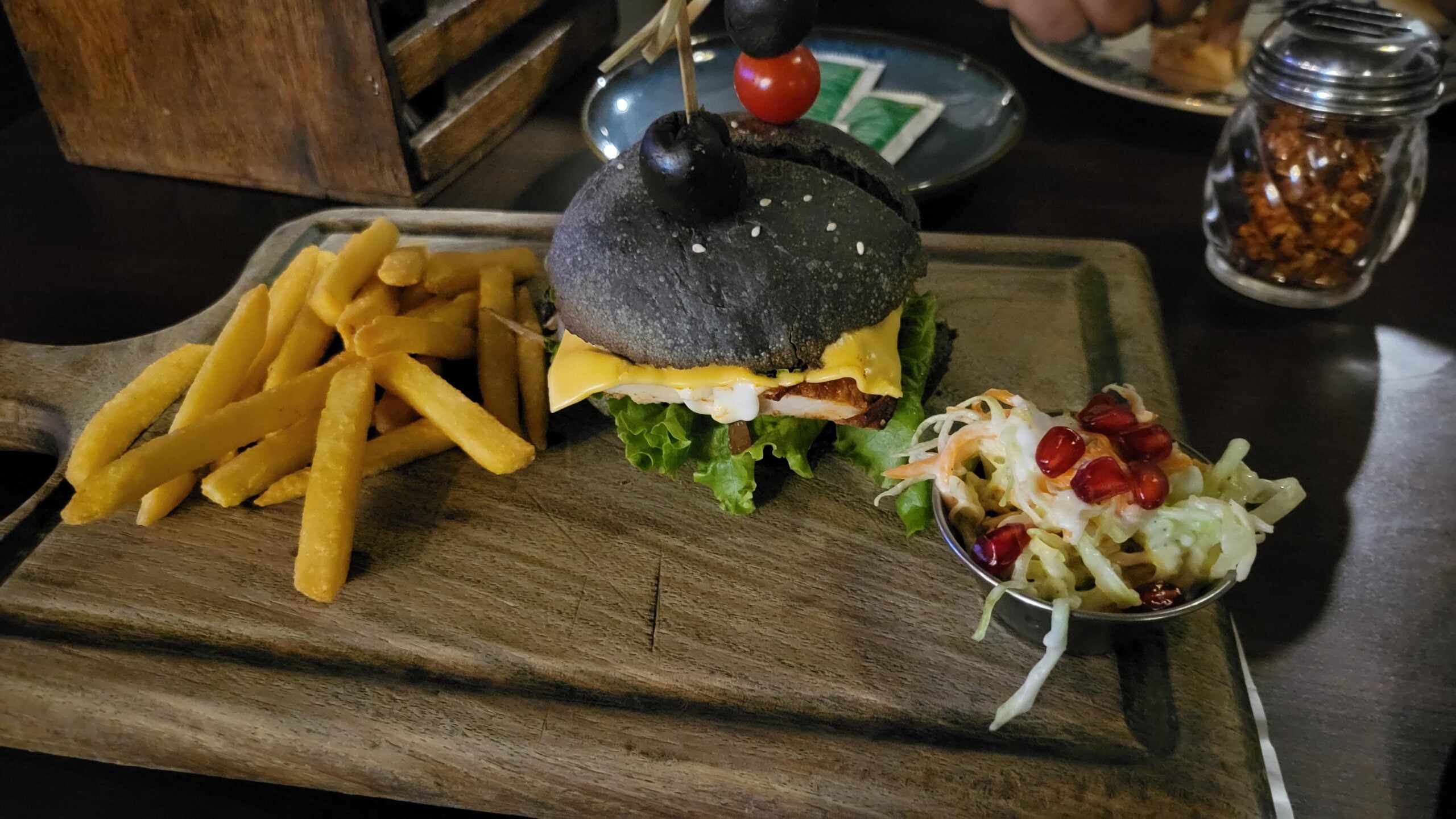 A charcoal burger with cheese, lettuce, and olives on top, served with French fries and a side of salad with pomegranate seeds on a wooden platter.