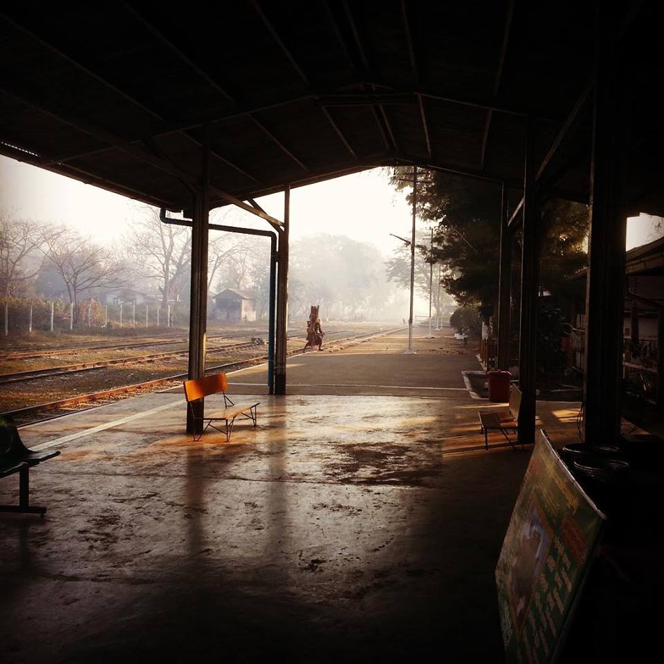 An empty railway platform with benches, bathed in soft morning light. Train tracks run alongside, trees and a small shed are visible in the background, creating a quiet, peaceful atmosphere.