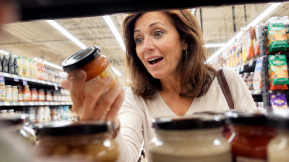 A woman in a grocery store looks excitedly at a jar she is holding, surrounded by other jars on the shelf, as she searches for the best store brand products.