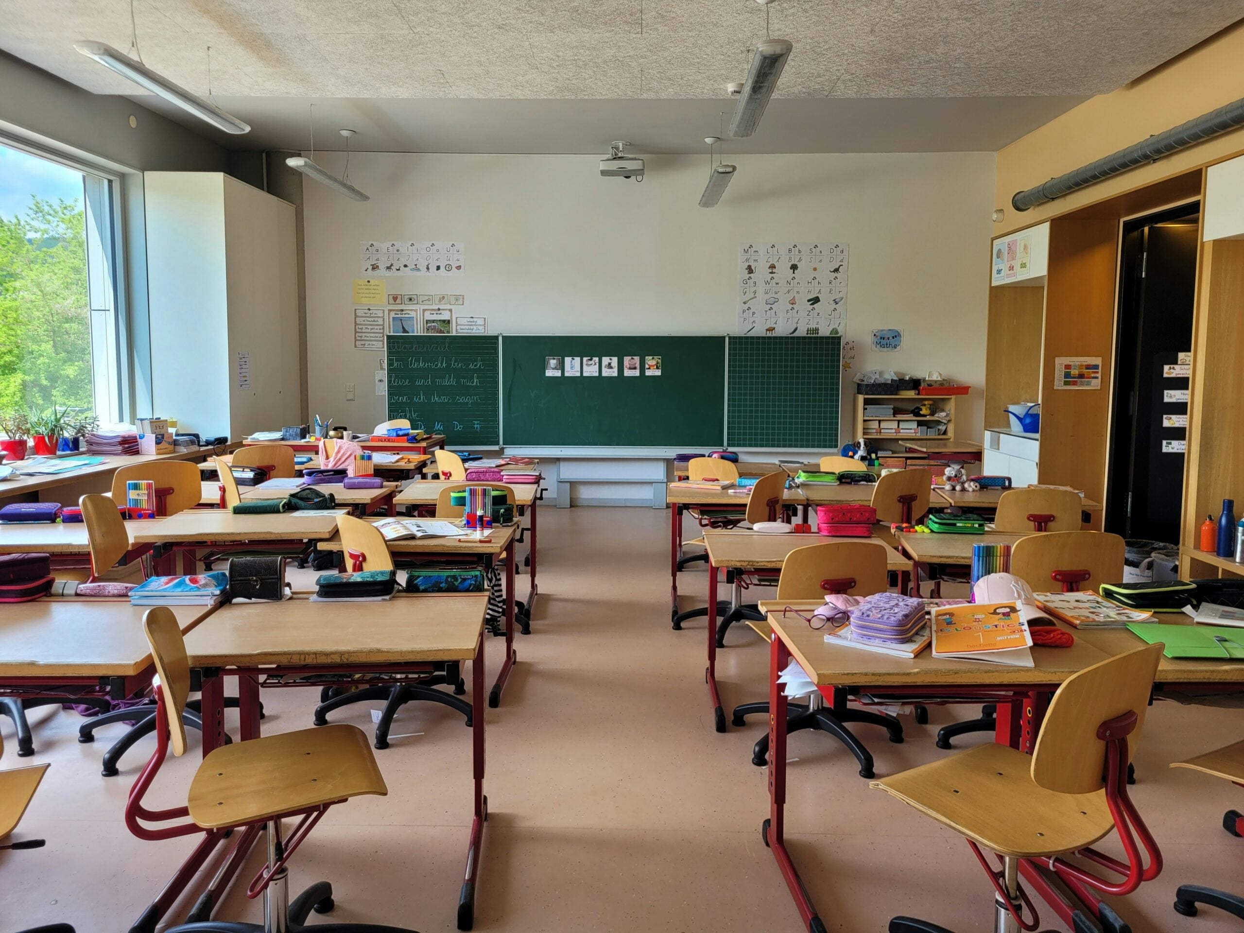 A classroom with wooden desks and chairs, scattered school supplies, and a large chalkboard at the front reflects the daily rhythm of life in 2026. Posters and drawings decorate the walls as sunlight streams in from windows on the left, offering a glimpse into how students adapt to new challenges and problems unique to their time.