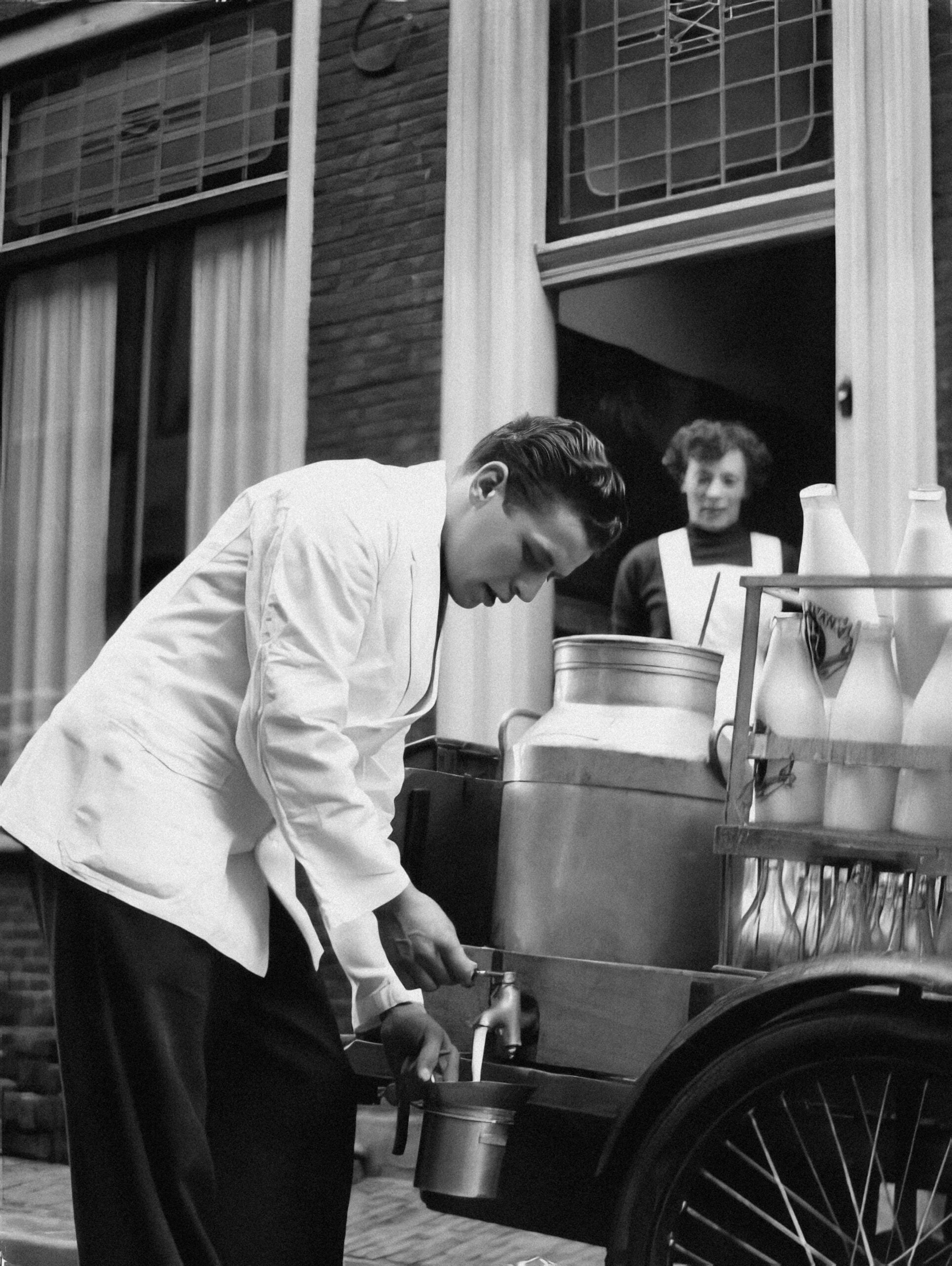 A man in a white coat fills a container from a milk cart outside a building, while a woman stands in the doorway watching him. Milk bottles and large metal containers are visible on the cart.