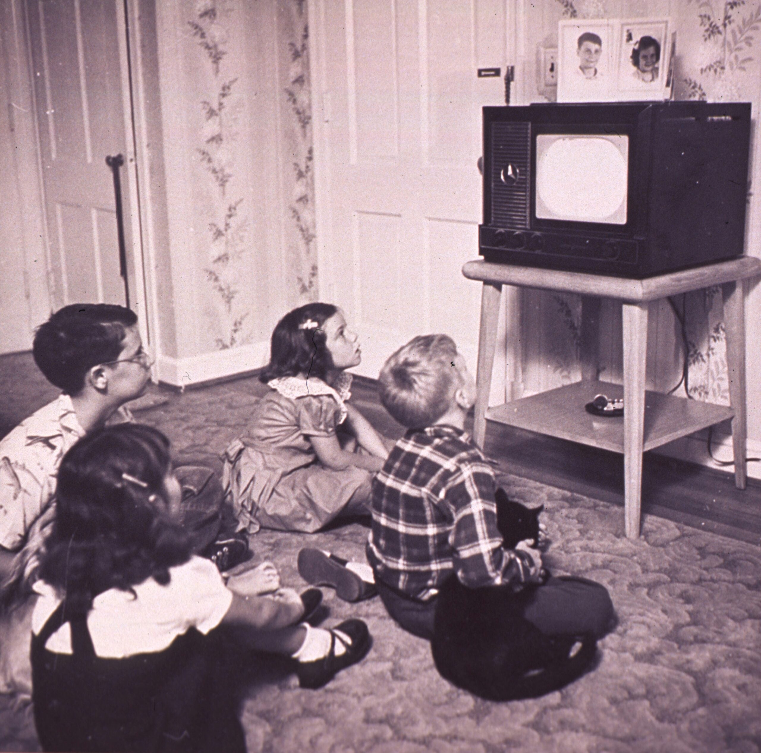 Four children sit on the carpet watching a small vintage television in a living room. Family photos rest on top of the TV, and a cat is on the boy’s lap. The scene has a nostalgic, 1950s atmosphere.