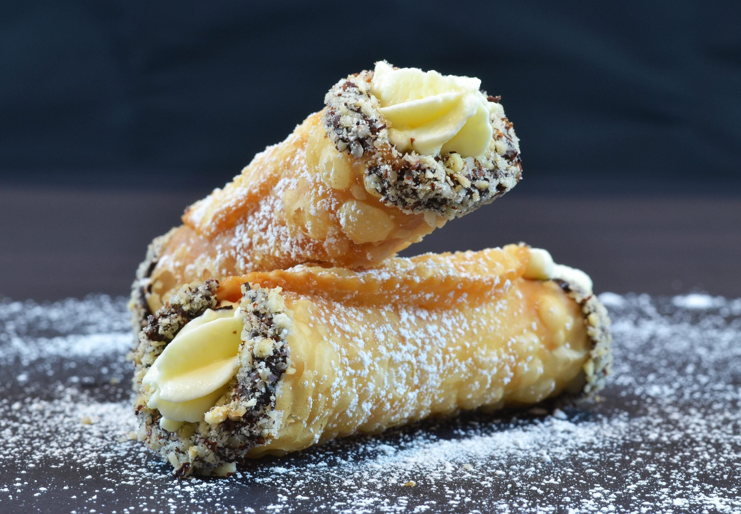 Two homemade cannolis filled with creamy filling, ends dipped in crushed nuts, stacked on a dark surface dusted with powdered sugar, against a black background.