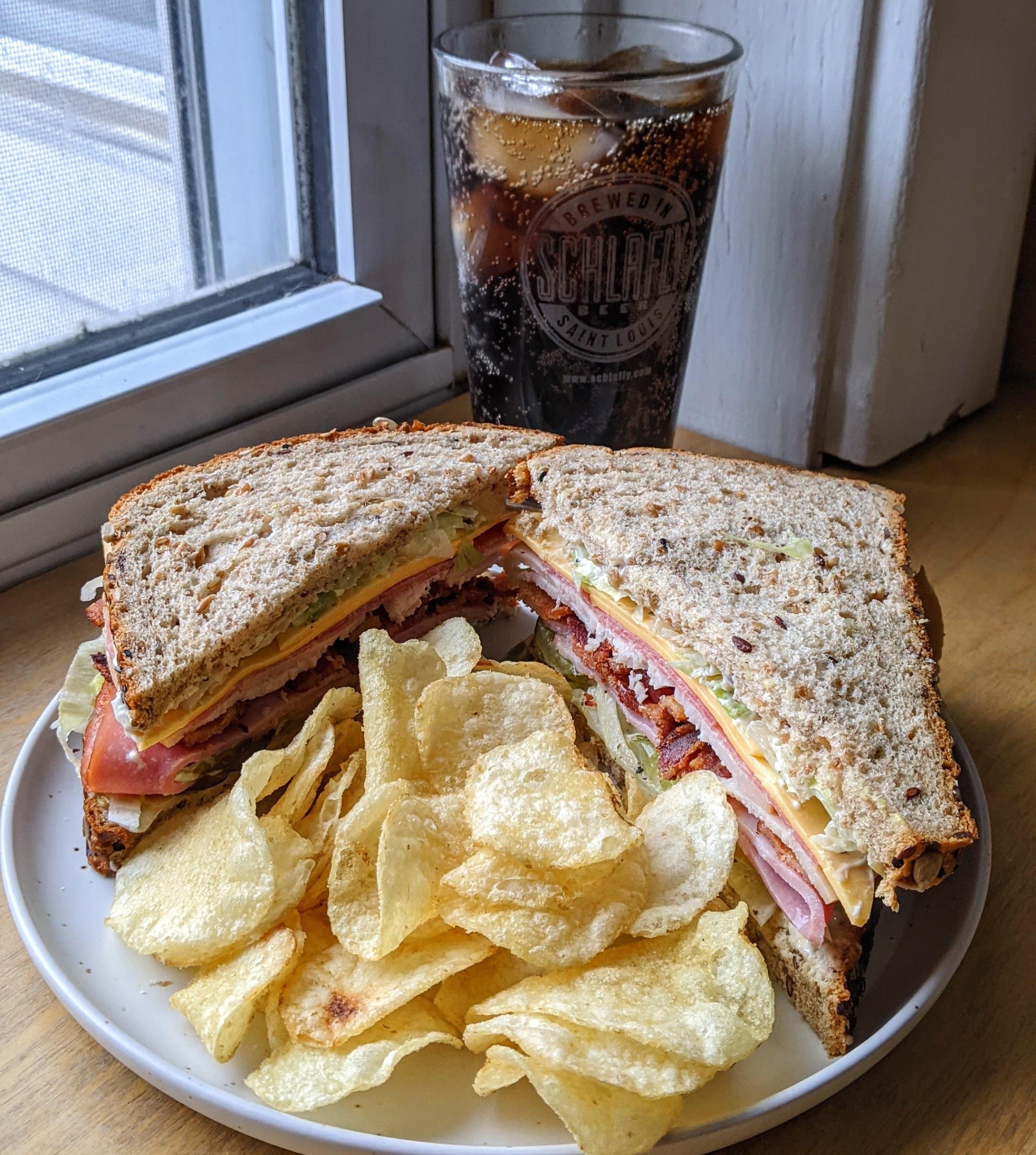 A plate with a deli sandwich cut in half, stacked with ham, cheese, lettuce, tomato, and bacon, served with potato chips. A glass of dark soda with ice sits behind the plate near a window.