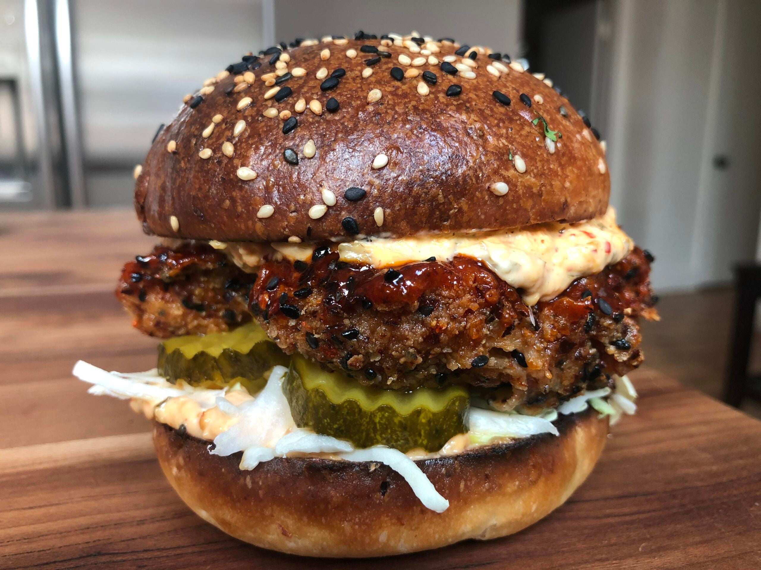 A close-up of a chicken sandwich with sesame seed bun, crispy chicken patty, pickles, shredded lettuce, and a creamy sauce, set on a wooden table.