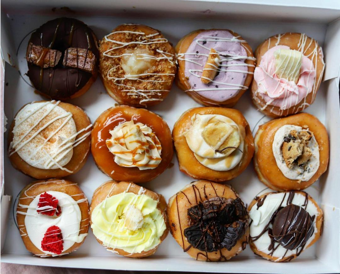 A box of twelve assorted donuts, each topped with colorful icing, drizzles, and various toppings like fruit, cookies, candies, and whipped cream.