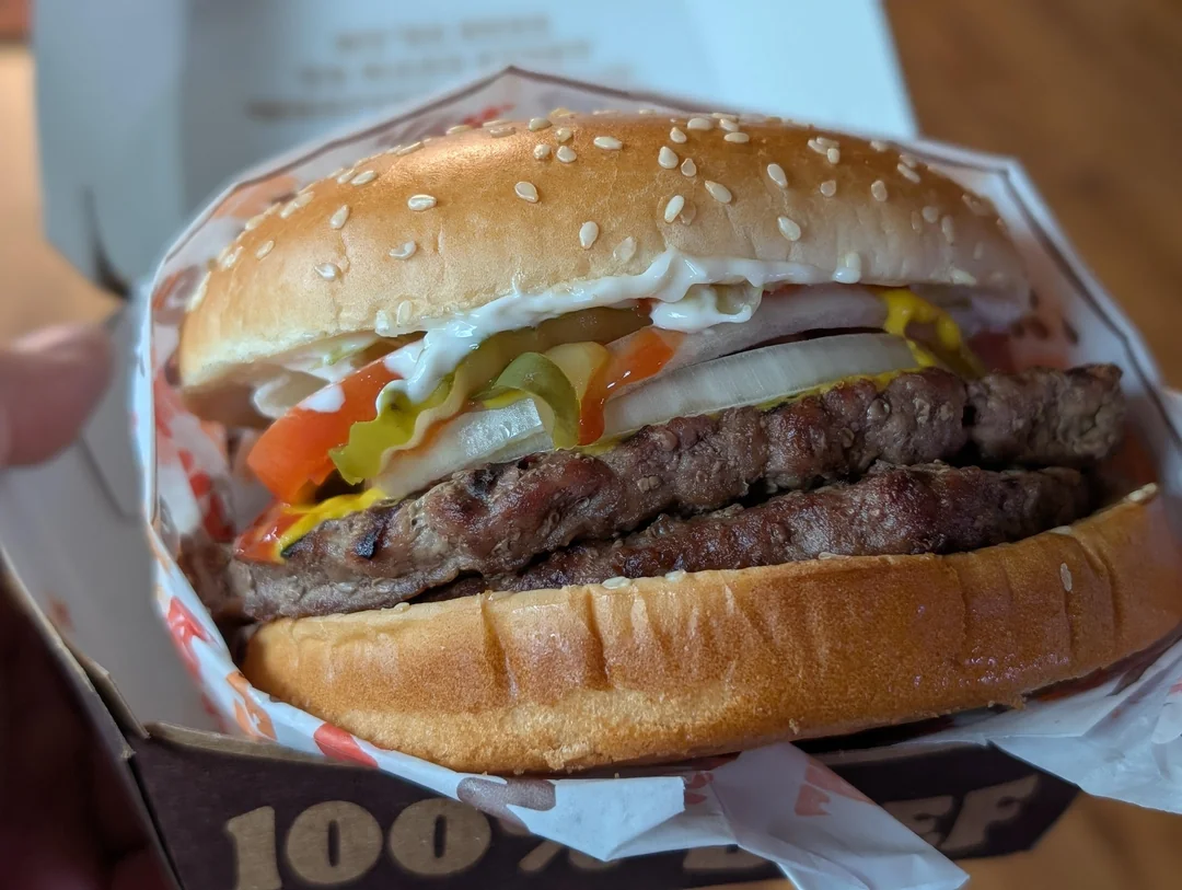 A close-up of a double cheeseburger with lettuce, tomato, onion, pickles, mayonnaise, and a sesame seed bun in a takeout box.