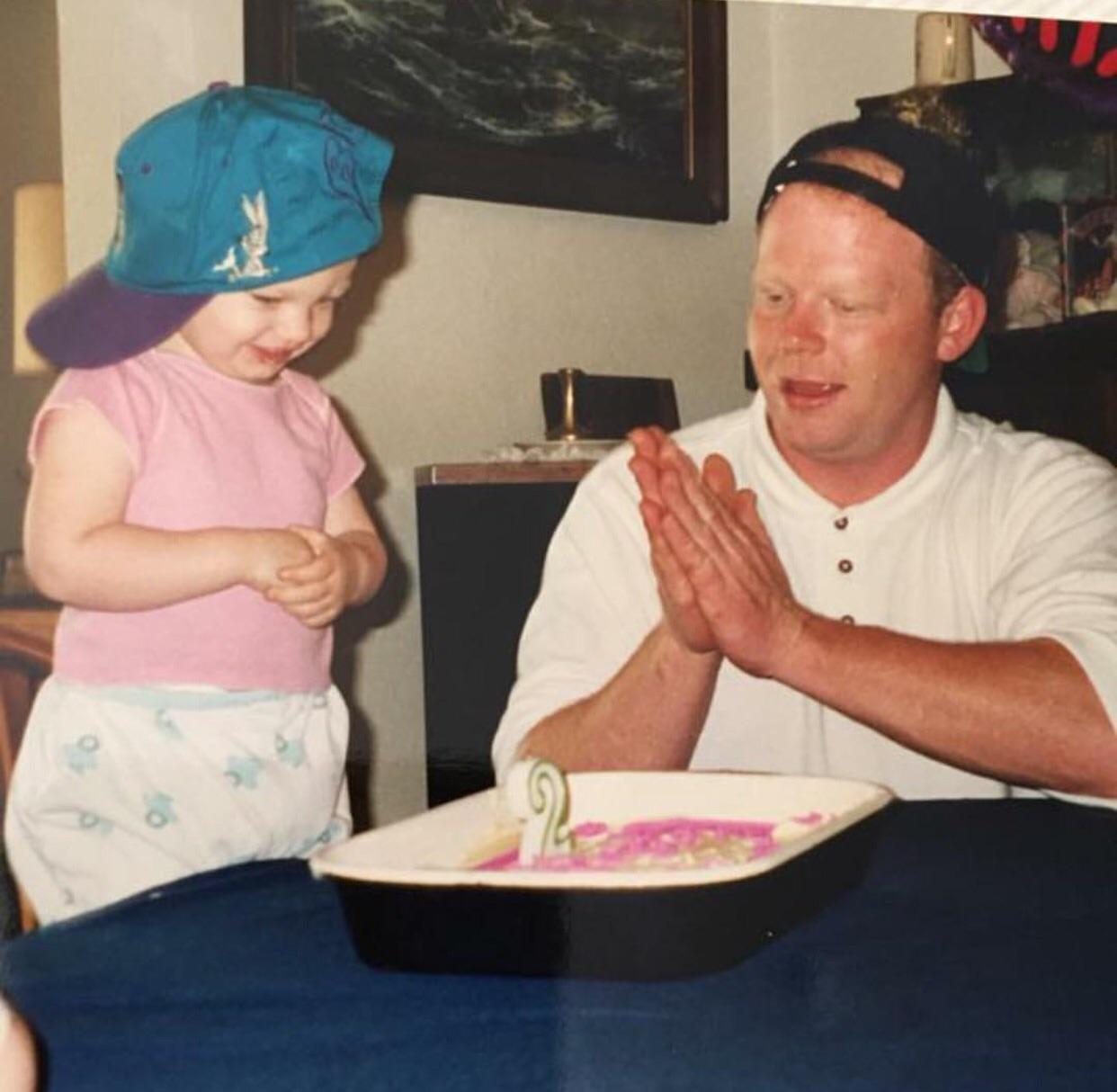 An adult and child wearing backward caps sit at a table with a cake that has a number 2 candle. The adult is clapping, and the child is smiling, looking excited.