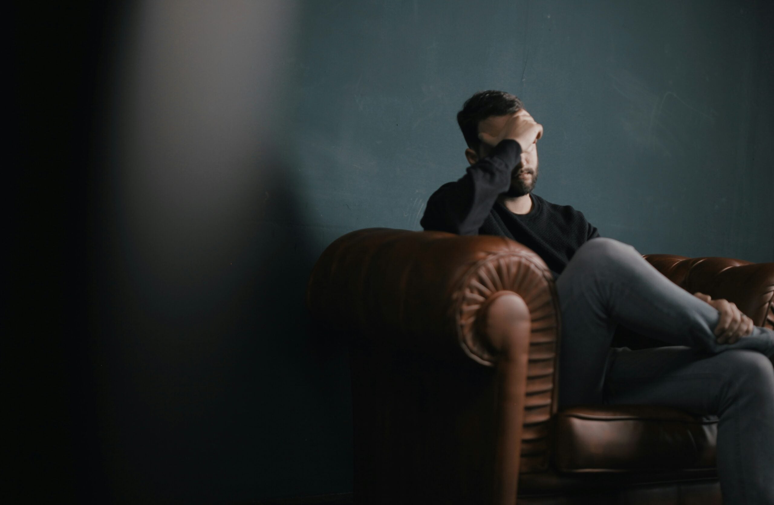 A man sits on a brown leather chair, resting his elbow on the armrest and covering part of his face with his hand, appearing stressed or deep in thought. The background is dark and blurred, reflecting the weight of life in 2026 problems on his mind.