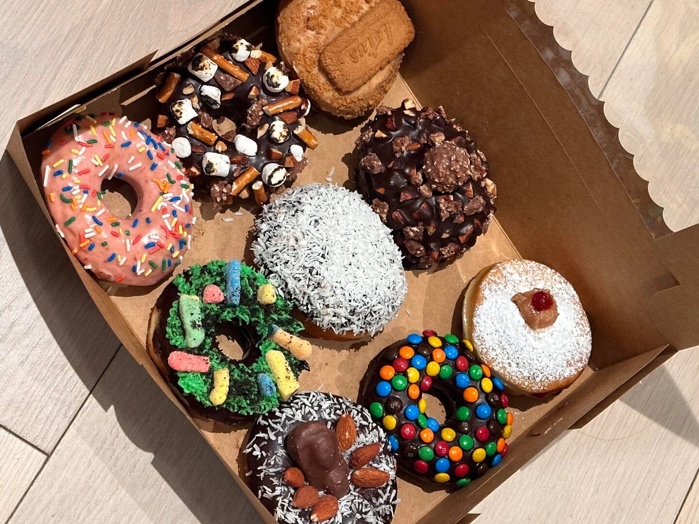 A box of nine assorted decorated donuts, each topped with colorful candies, chocolate, coconut flakes, sprinkles, cookies, or marshmallows, sits open on a light wooden surface in bright sunlight.