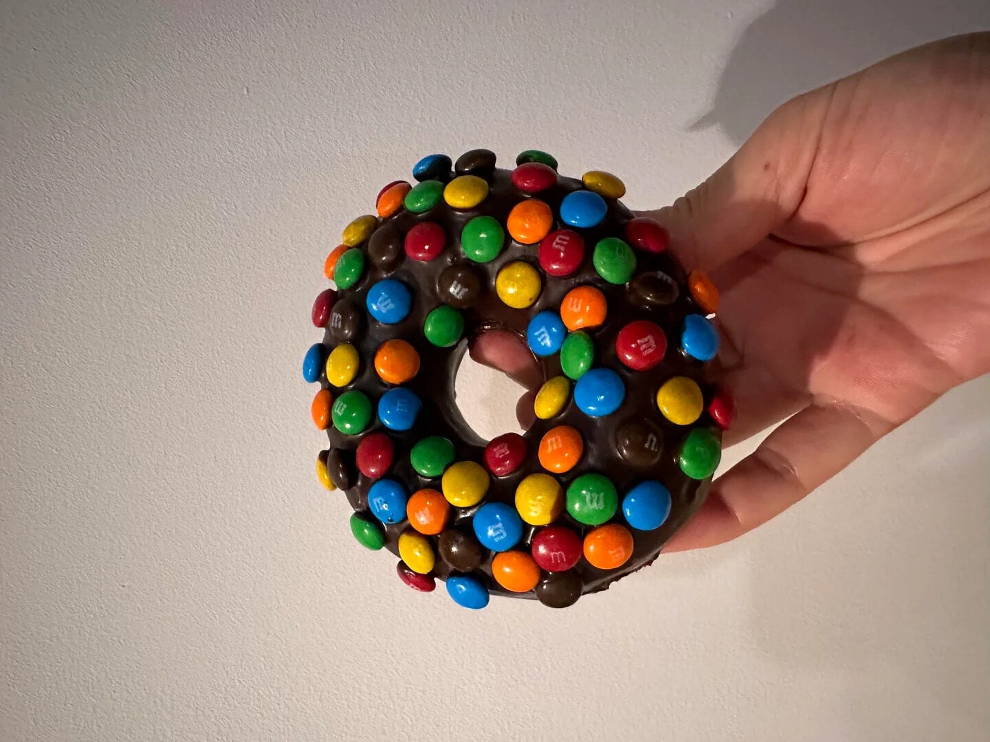 A hand holds a chocolate-frosted donut covered with colorful M&M candies against a plain, light-colored background.