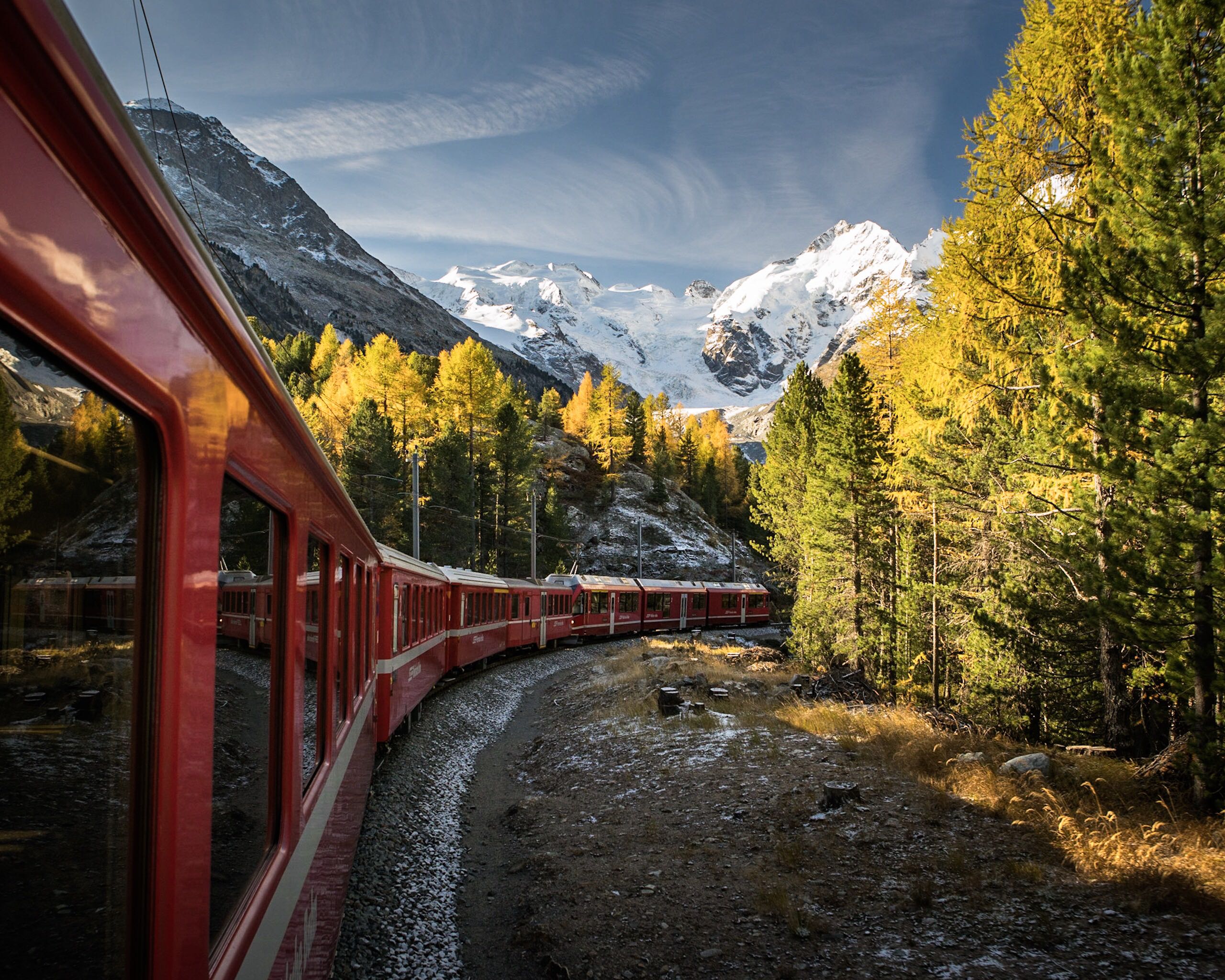 A red train winds through a forest with yellow autumn trees, heading toward snow-capped mountains under a blue sky with wispy clouds.