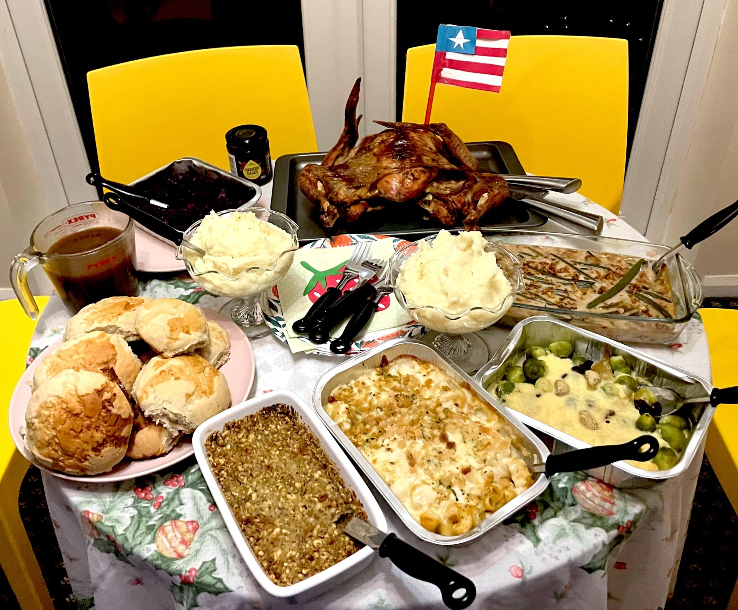 A table set with roast meat, mashed potatoes, bread rolls, green bean casserole, macaroni and cheese, Brussels sprouts, and a Liberian flag, all arranged on a floral tablecloth with yellow chairs.