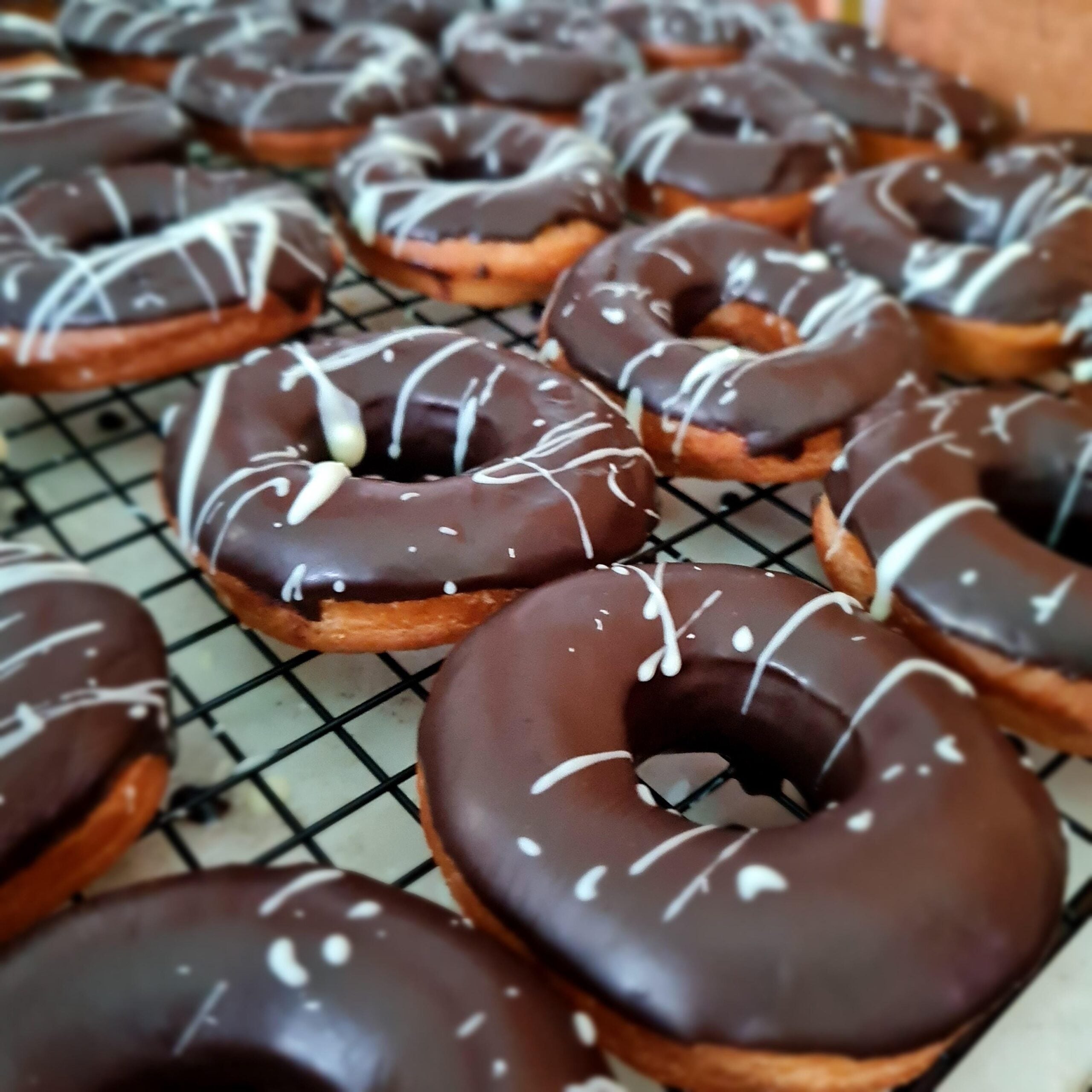 Chocolate-glazed donuts with white icing drizzle are arranged on a cooling rack. The donuts are golden brown and the chocolate glaze looks shiny and smooth. The background is slightly out of focus.