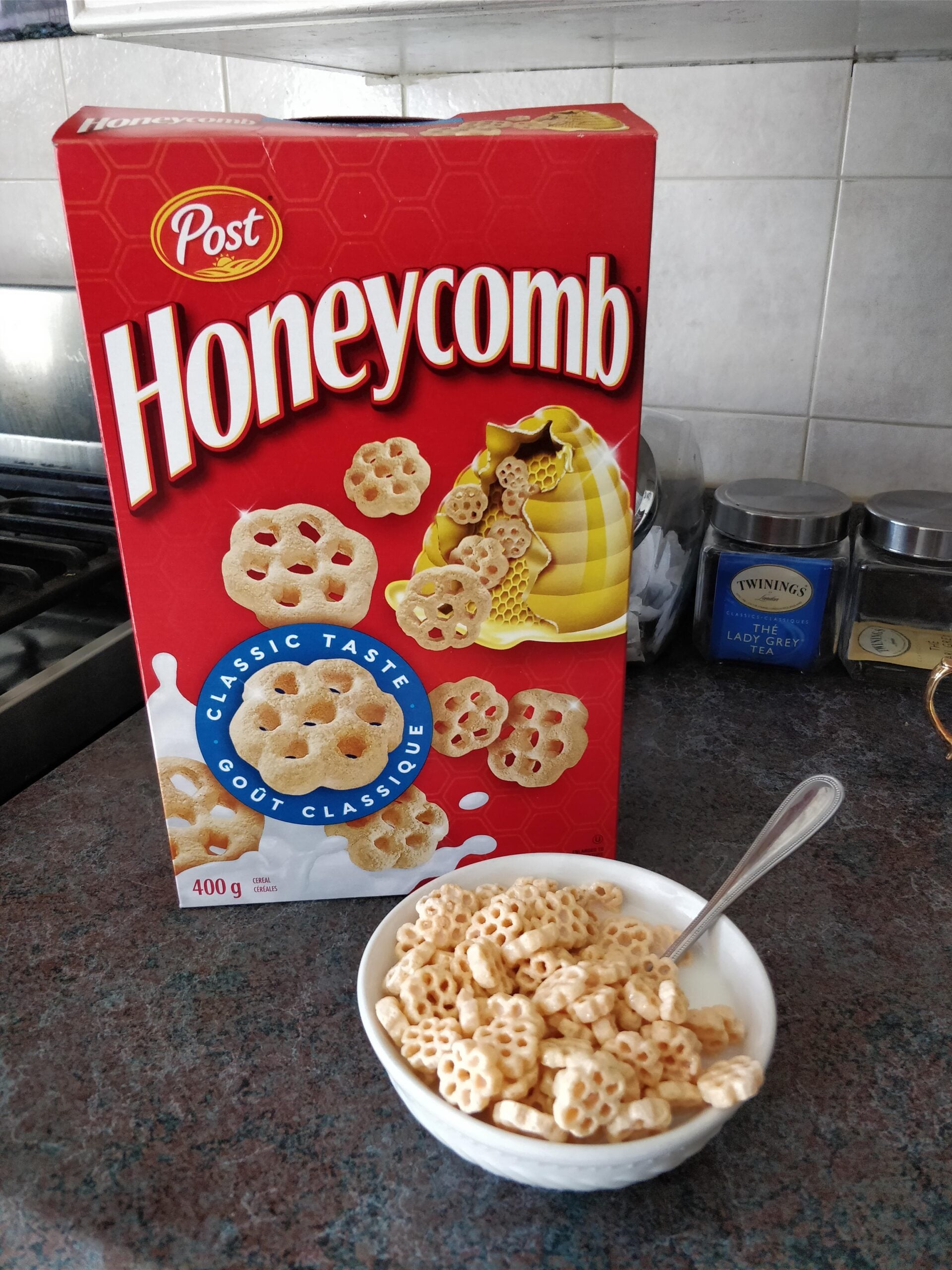 A box of Post Honeycomb cereal is on a kitchen counter next to a bowl filled with the cereal and a spoon. Tea tins and a kettle are in the background.