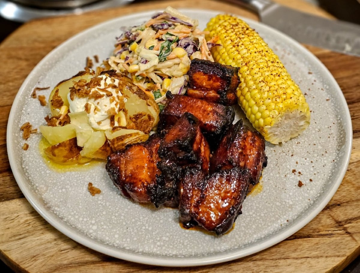 A plate with glazed barbecue ribs, corn on the cob, a baked potato with sour cream and onions, and creamy coleslaw on a light grey dish.