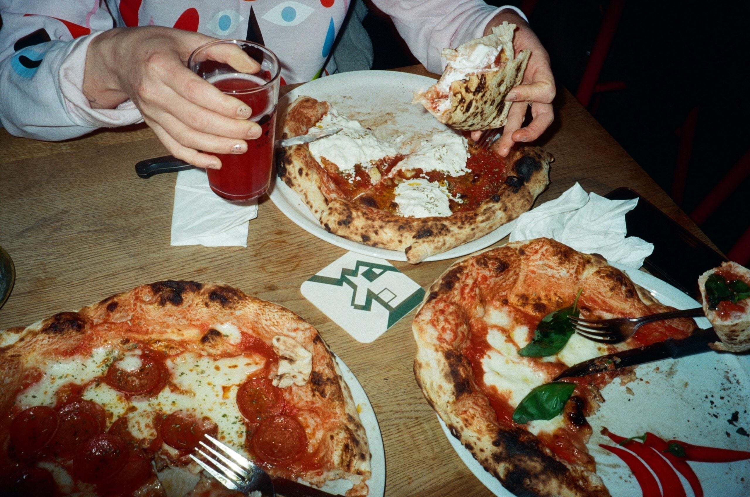 Three pizzas with different toppings are on a wooden table. A person in a colorful shirt holds a slice of pizza in one hand and a glass of red drink in the other. Plates and napkins are scattered around.
