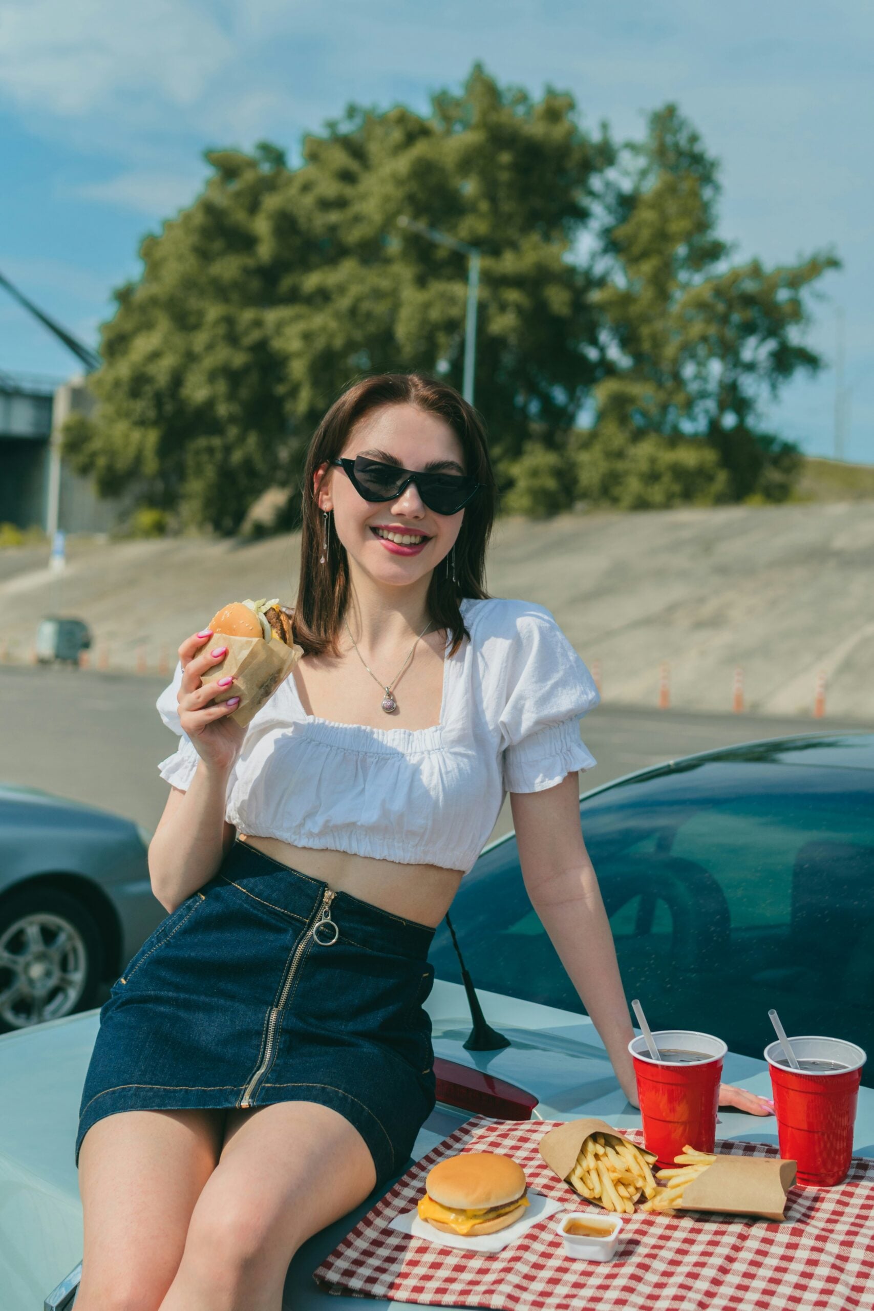 A woman in sunglasses and a white crop top sits on a car eating a burger. Fries, burgers, and drinks are on a red-checkered cloth on the car. It’s sunny and there are trees in the background.