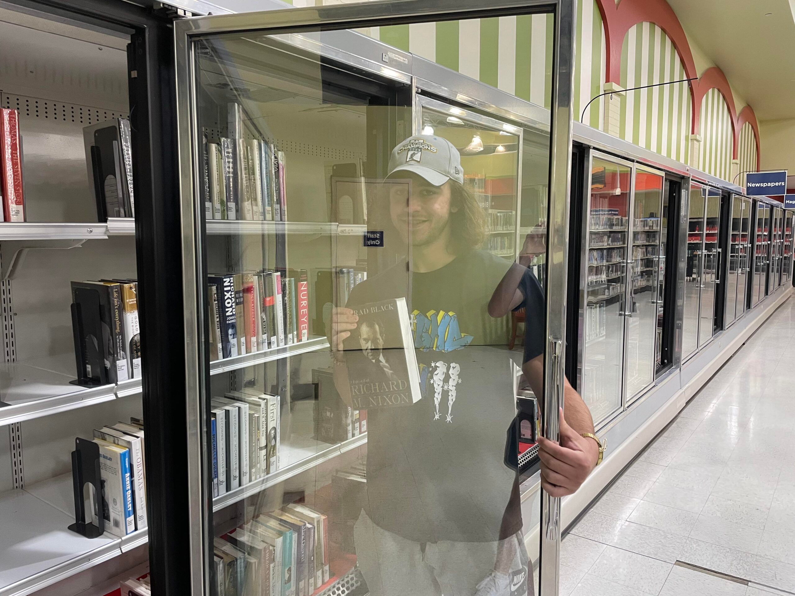 A person stands inside an open grocery store refrigerator filled with books, holding a book and smiling. The scene combines elements of a supermarket and a library.
