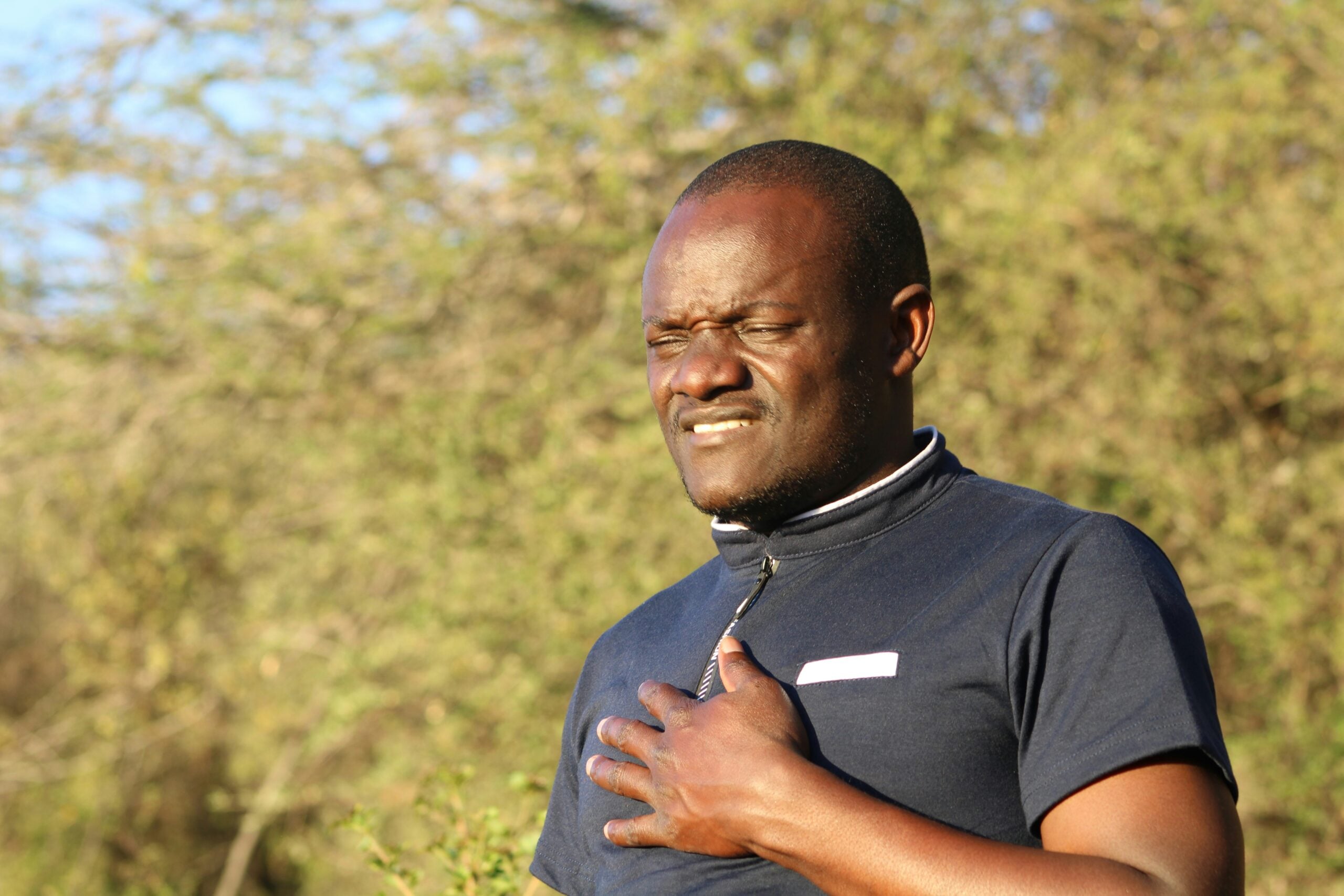 A man stands outdoors, eyes closed and expression tense, holding his hand to his chest as if in discomfort or pain—perhaps weighed down by the pressures of life in 2026. Green foliage is visible in the background.