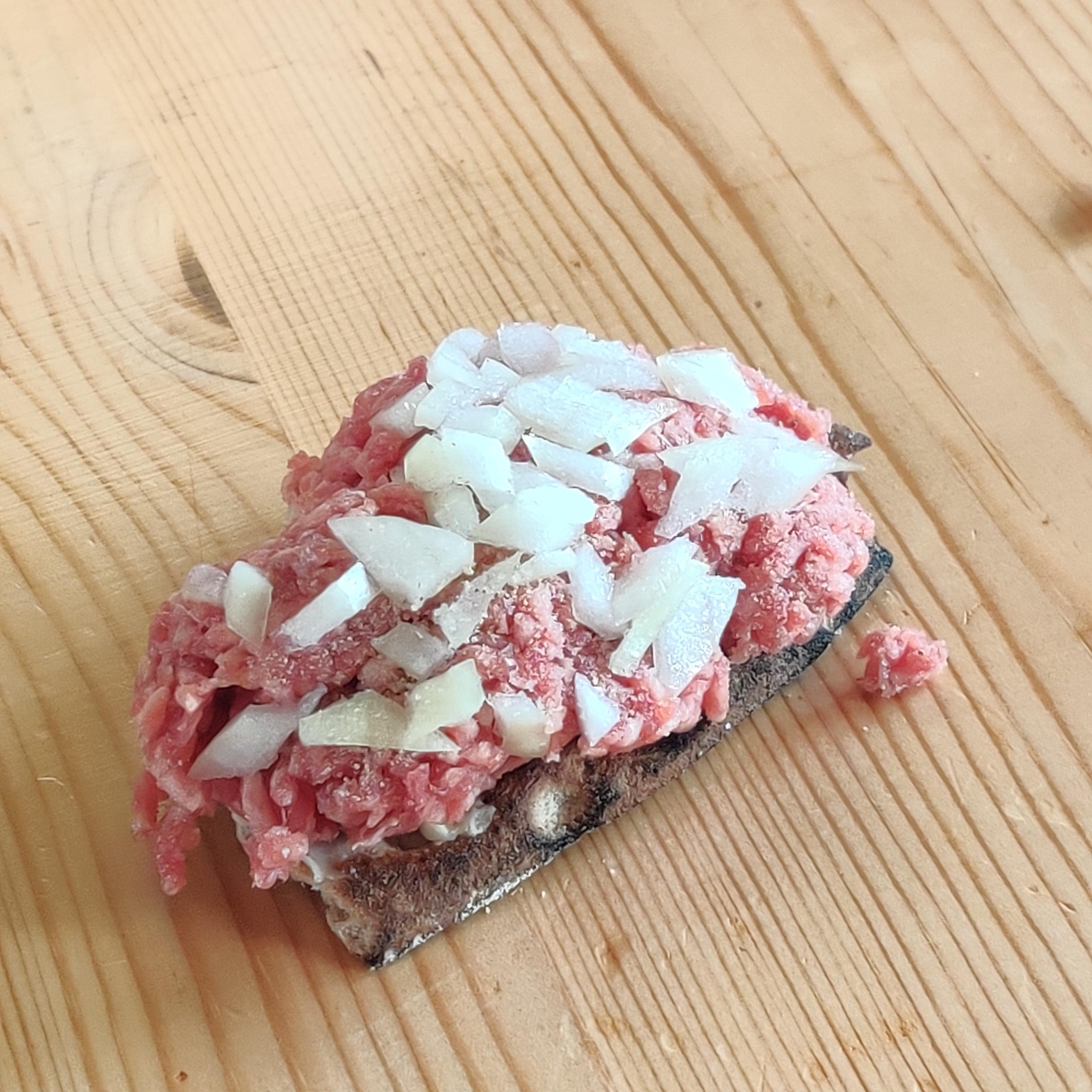 A slice of dark bread topped with raw ground meat and chopped white onions sits on a light wooden surface.