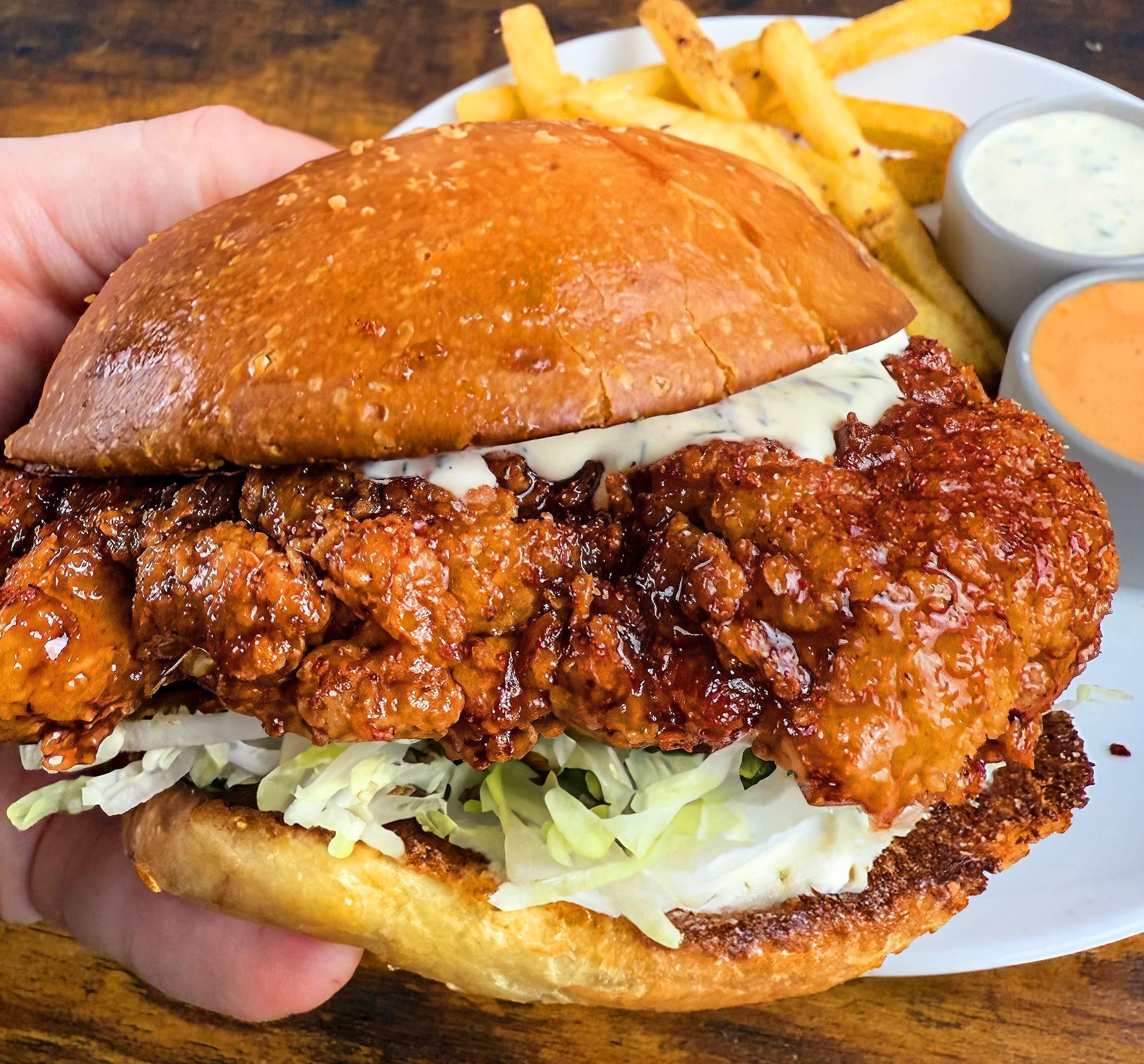 A hand holds a crispy fried chicken sandwich with lettuce and sauce on a toasted bun, served with fries and two dipping sauces on a white plate.