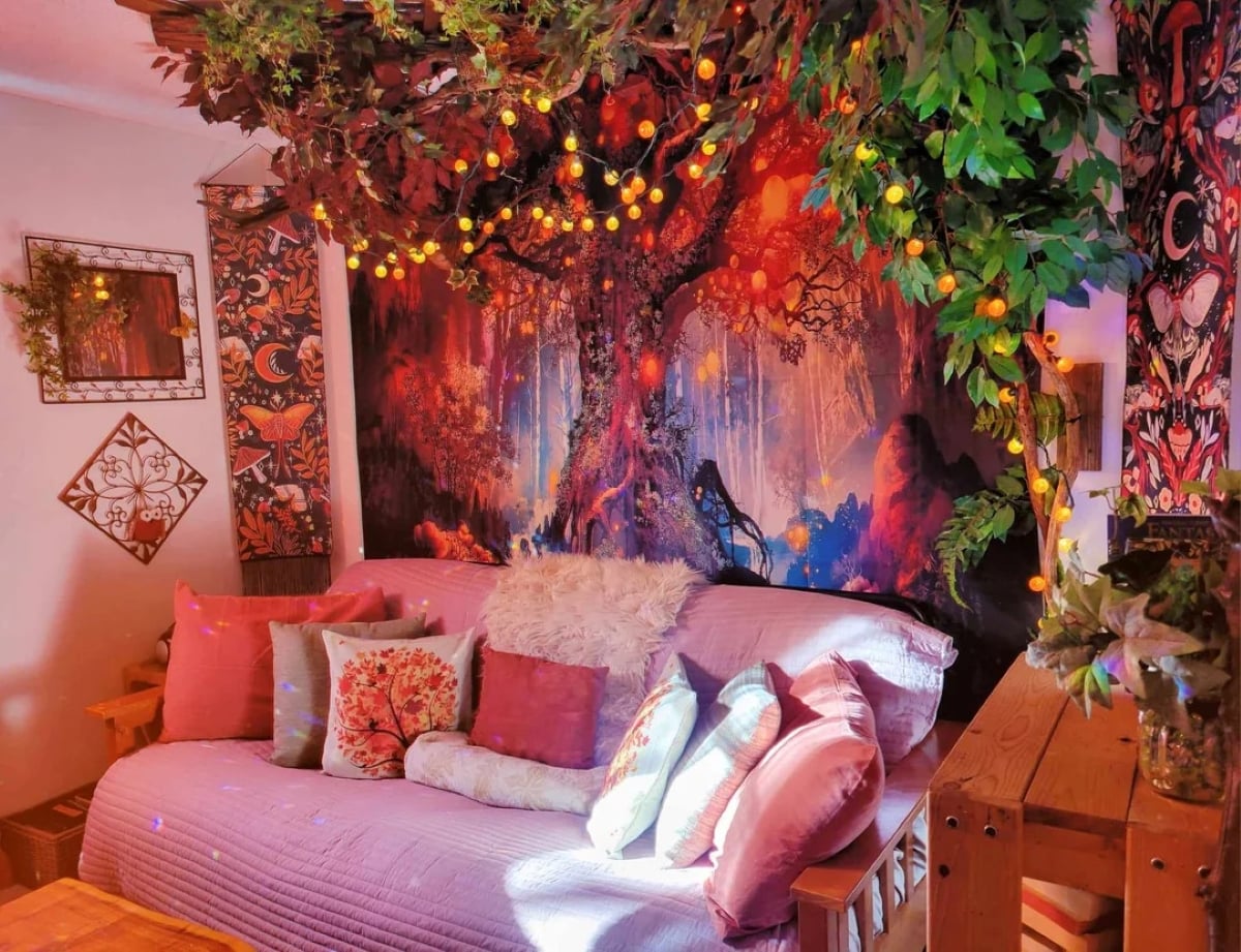 A cozy room with a couch covered in pink and floral pillows, a large fantasy tree tapestry on the wall, and leafy garlands with glowing string lights hanging from the ceiling.