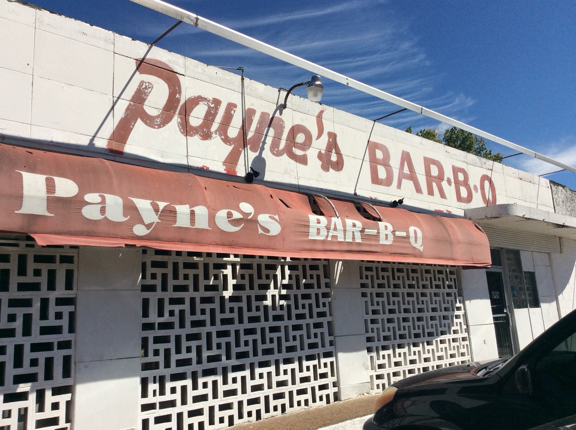 Payne’s BAR-B-Q restaurant exterior with a faded red sign and awning, white geometric brick wall, and a black car parked in front under a blue sky.