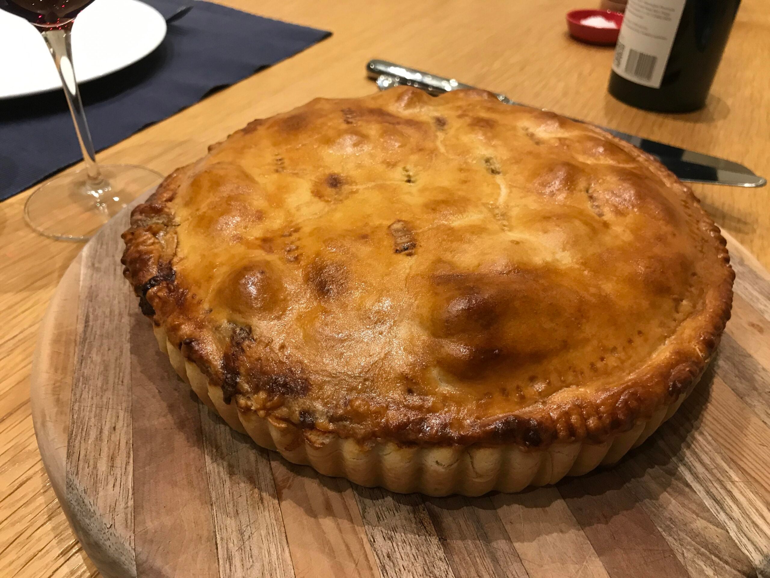 A golden-brown pie with a shiny crust sits on a wooden board, next to a glass of red wine, a bottle, and cutlery on a wooden table.
