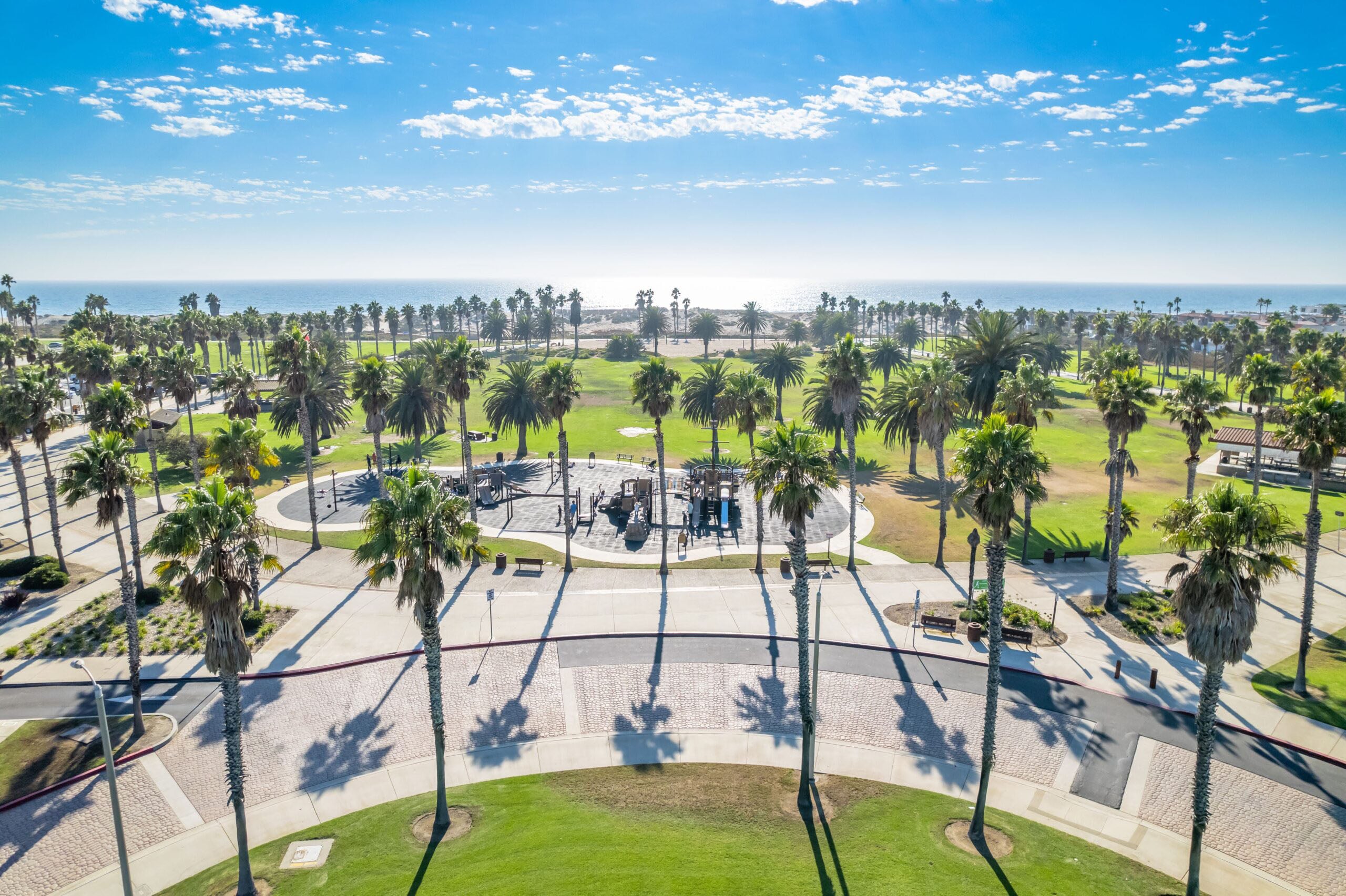 A sunny park with palm trees, a playground, green lawns, and walking paths, leading toward the ocean in the background under a blue sky with scattered clouds. Best cities to start your career.