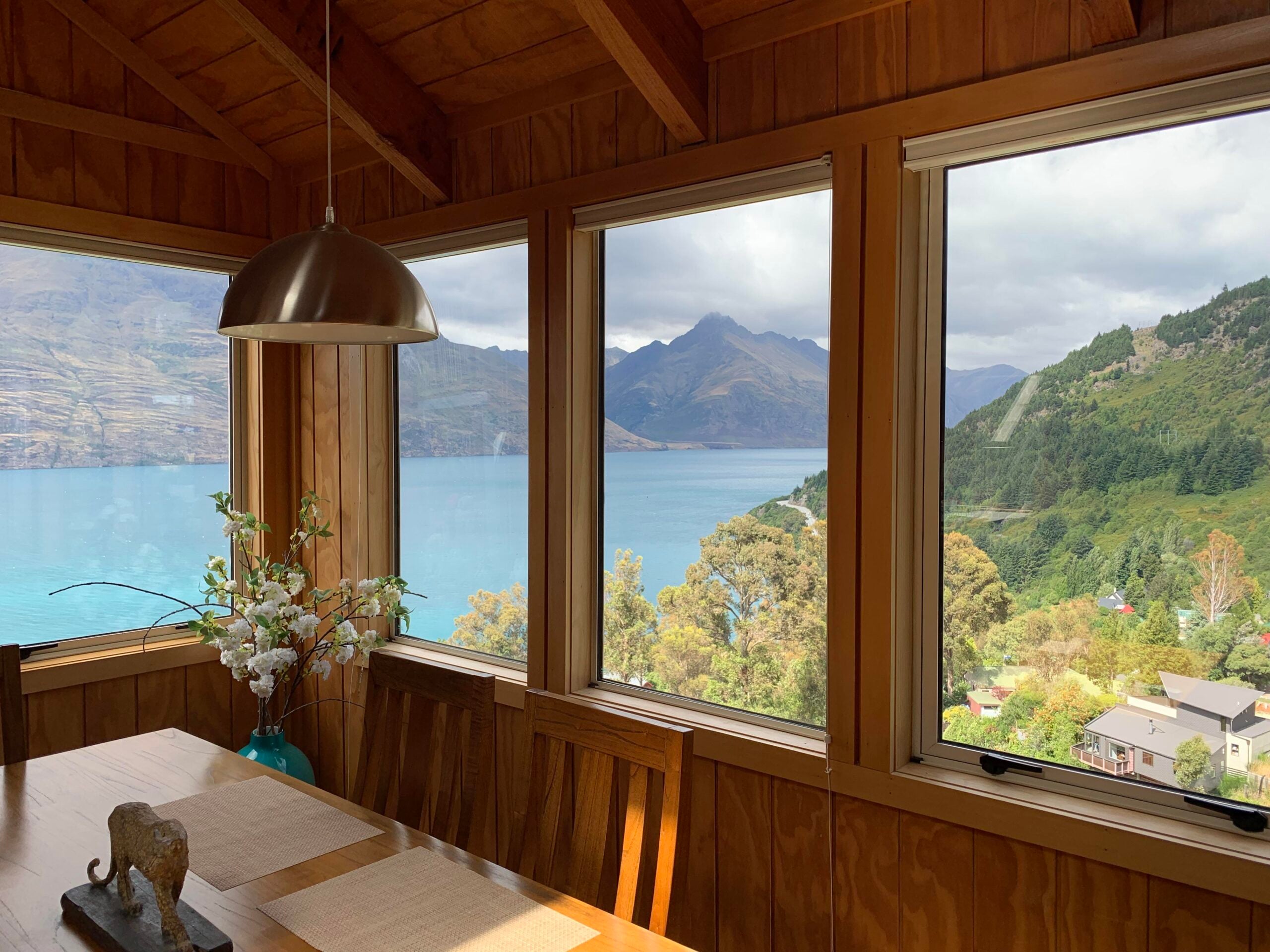 A cozy wooden dining area with large windows overlooks a blue lake, green hills, and distant mountains under a partly cloudy sky. A vase with flowers sits on the table.