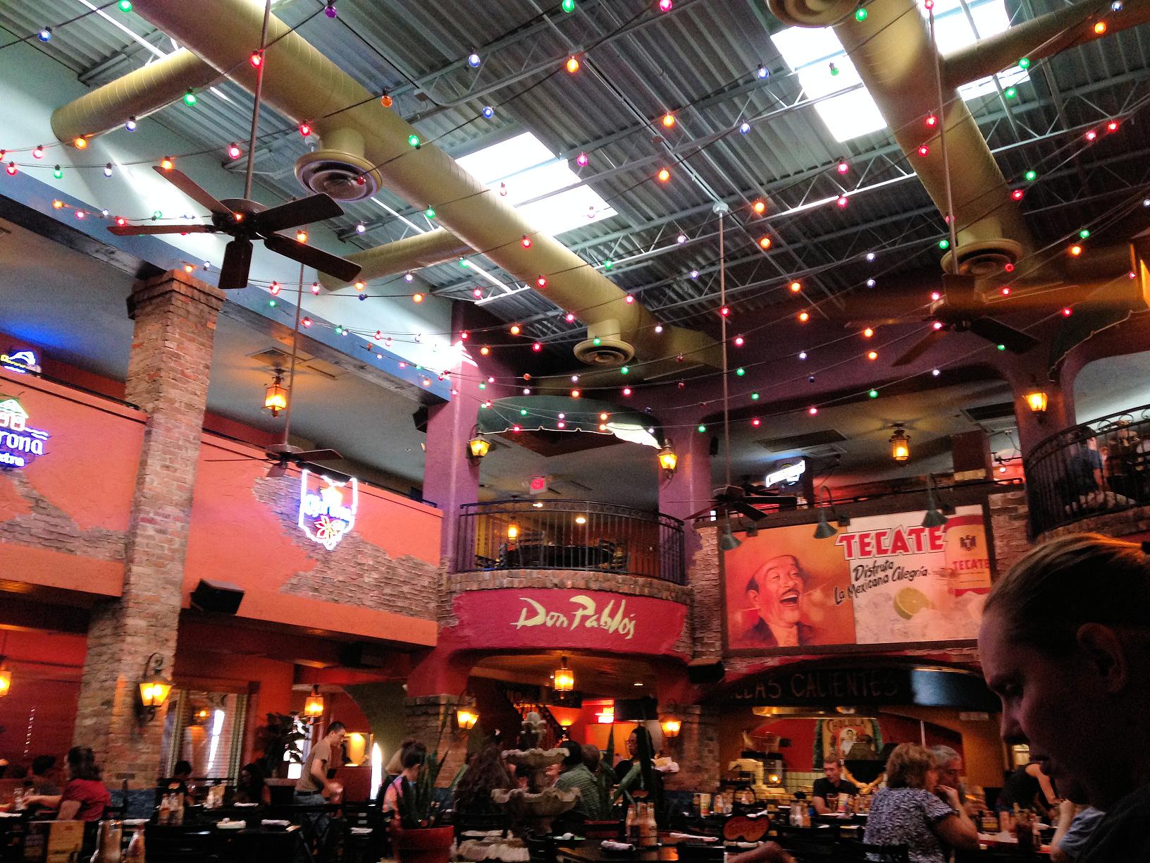 Interior of a lively Mexican restaurant with colorful string lights, rustic decor, brick columns, and diners seated at tables. Bright banners and advertisements decorate the upper walls.