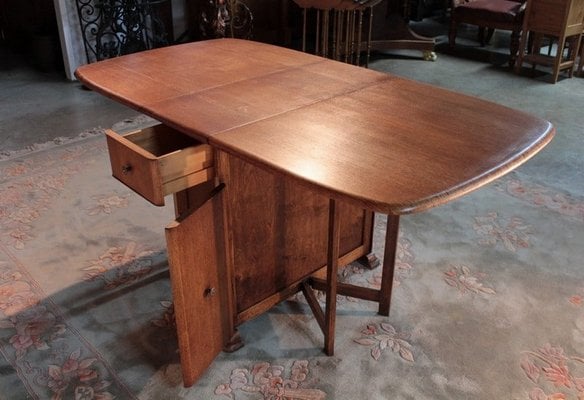 A wooden drop-leaf table with both leaves extended, one side featuring a small open drawer and a cabinet door, positioned on a patterned rug.