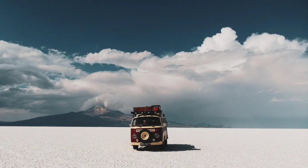 A vintage van with luggage on top sits alone on a vast white salt flat, evoking the spirit of classic road trips under a blue sky with scattered clouds and distant mountains.