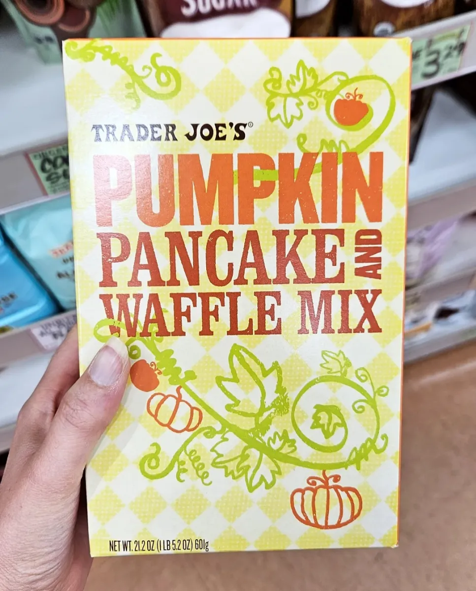A hand holds a box of Trader Joe’s Pumpkin Pancake and Waffle Mix with yellow and orange text and pumpkin illustrations on a checkered background.