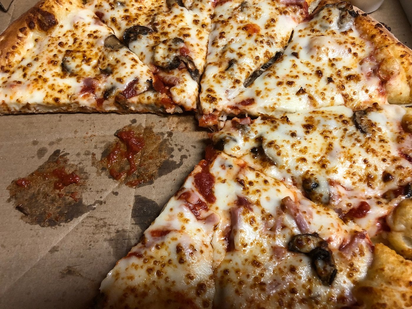 A pizza with melted cheese, mushrooms, and pieces of meat sits in an open pizza box. Two slices have been removed, leaving a gap, and some sauce is visible on the box where slices were taken.