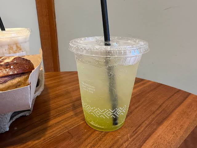 A clear plastic cup with a lid and black straw contains a light green, fizzy drink on a wooden table. A food container with baked goods is partially visible to the left. Restaurant chains with fall menu items.