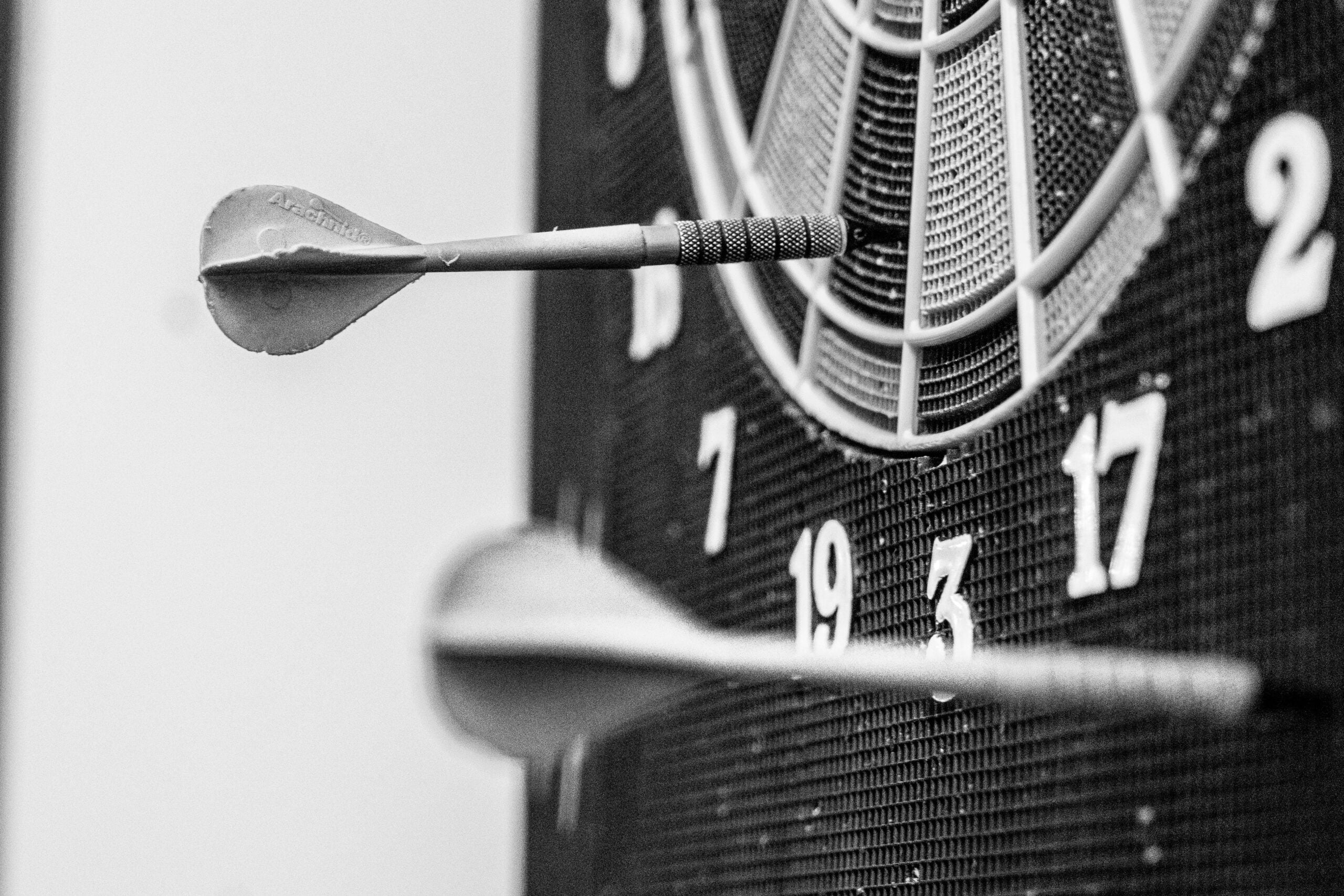 Black and white close-up of two darts stuck in a dartboard, one in the bullseye area and the other near the number 19. The image emphasizes focus and precision.