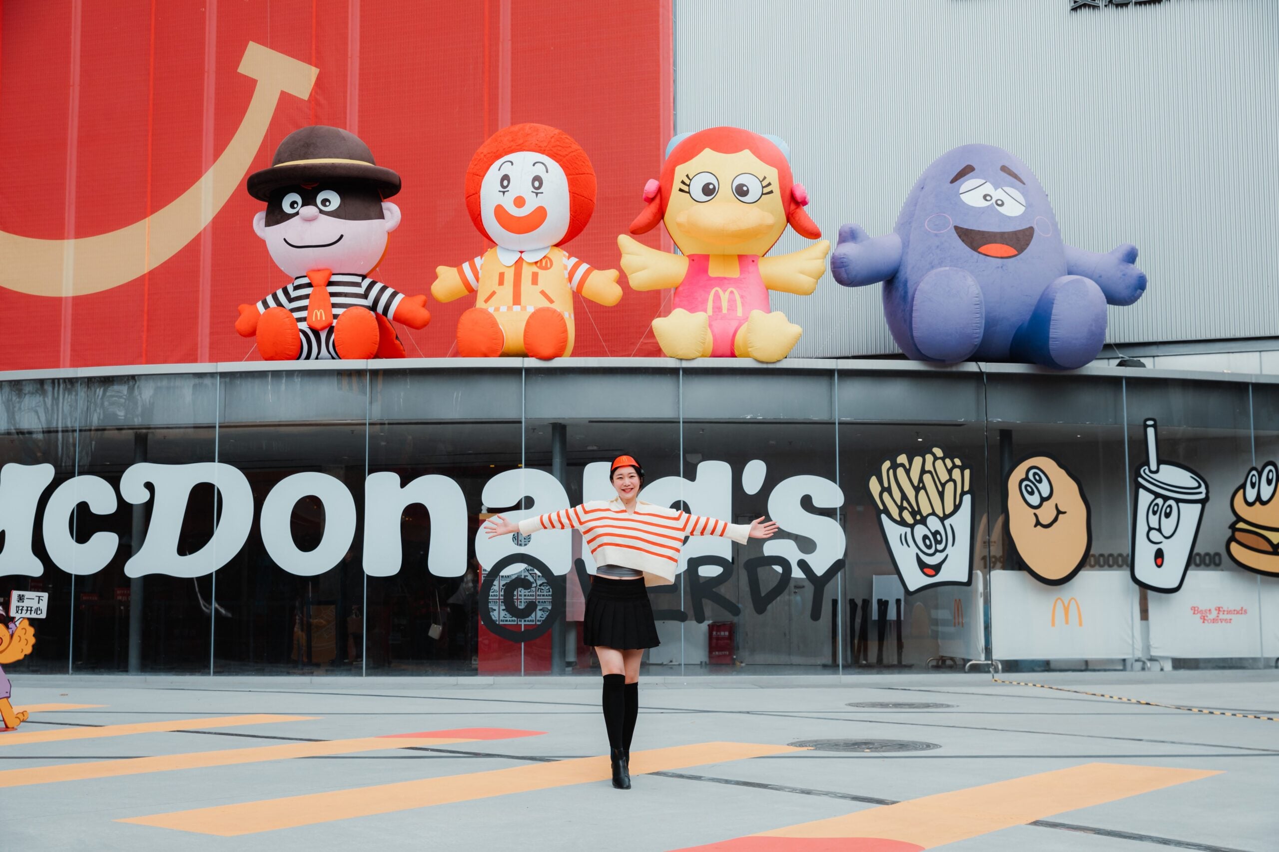 A woman in a striped shirt and black skirt poses with arms outstretched in front of a McDonald’s, with large character inflatables displayed above the entrance.