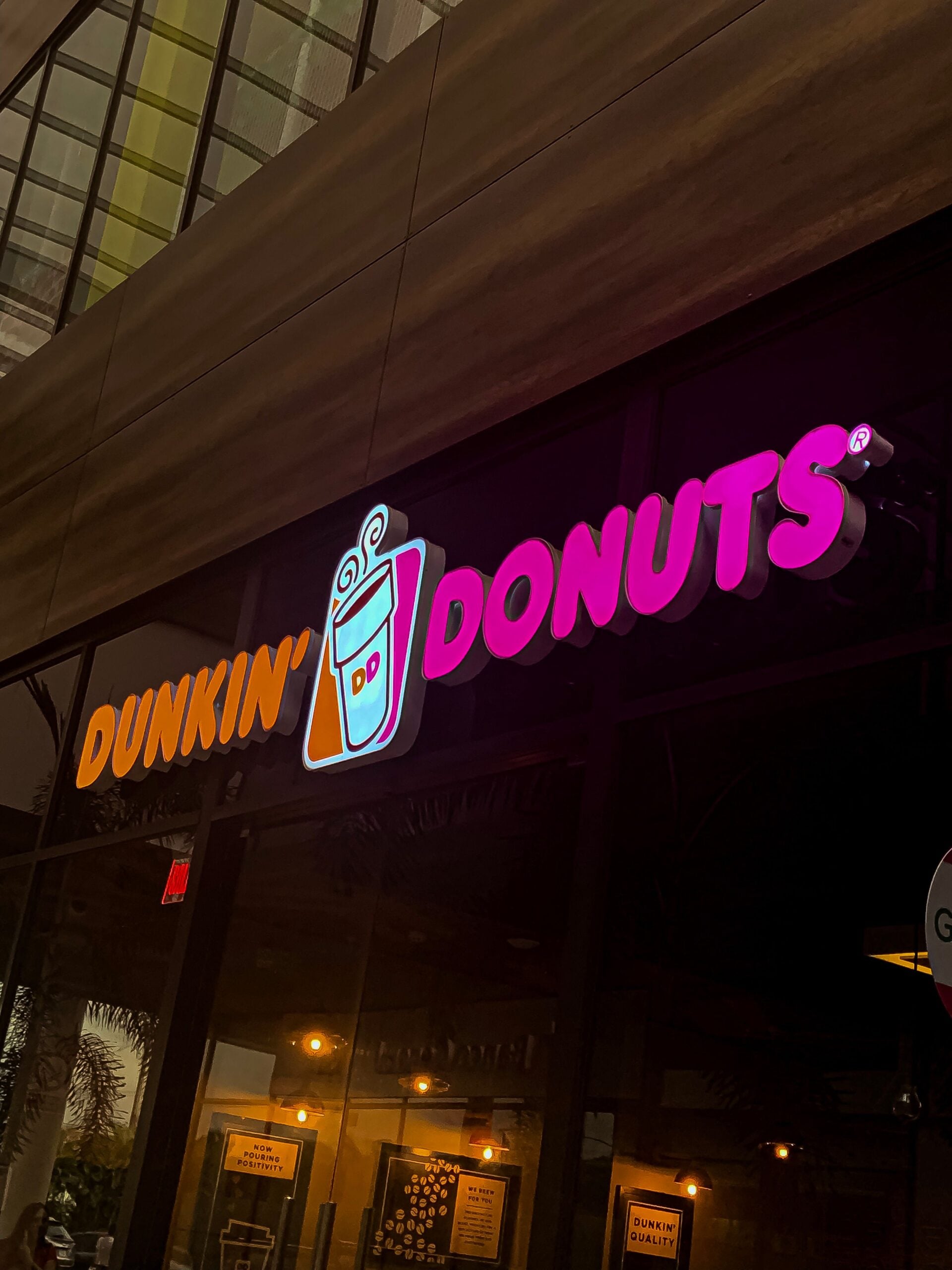 Dunkin’ Donuts storefront with illuminated pink and orange sign and coffee cup logo, seen at dusk with reflection of palm trees and building in the glass.