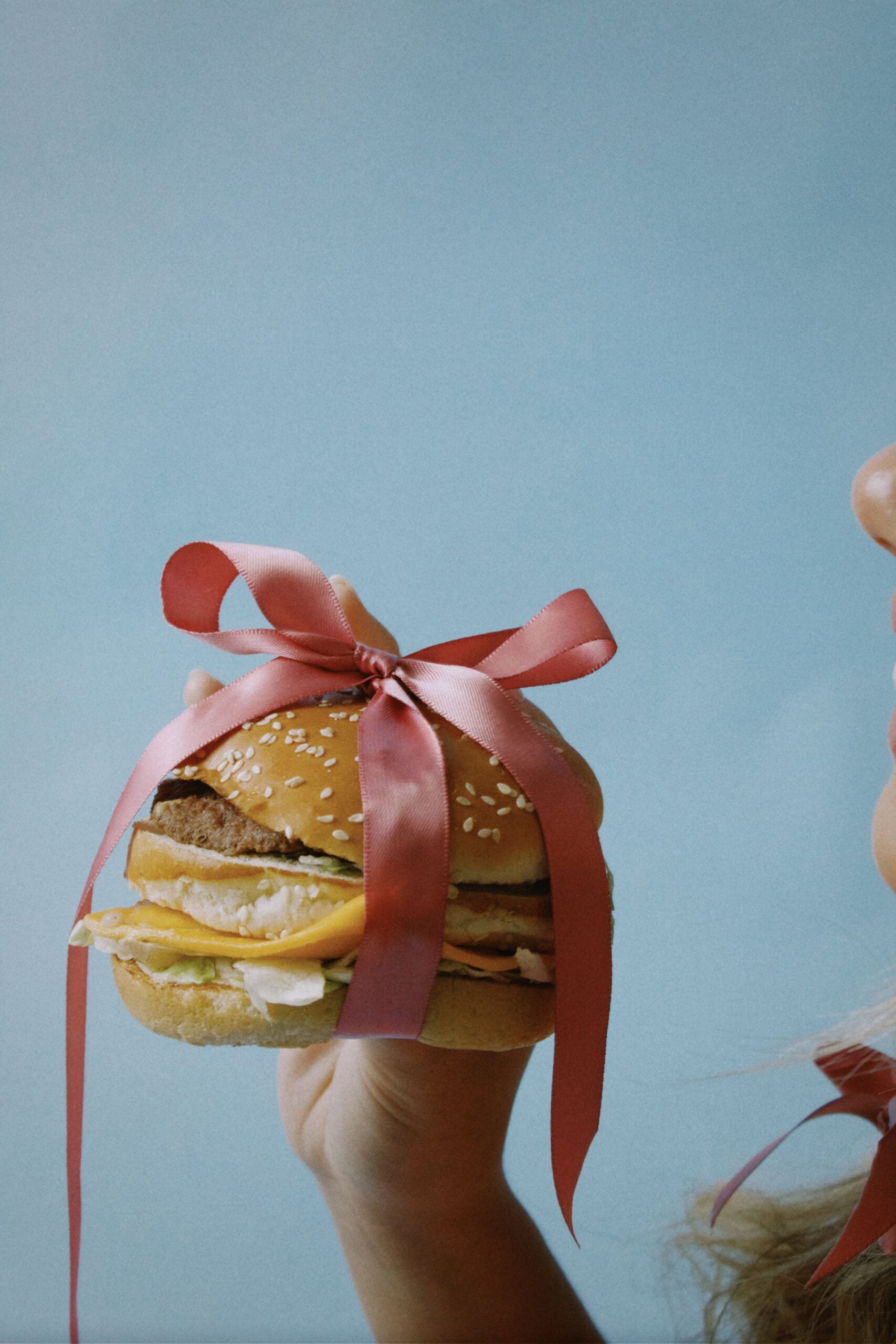 A hand holds a hamburger tied with a pink ribbon, as if giving it as a gift. A person’s face is partially visible on the right side against a light blue background.