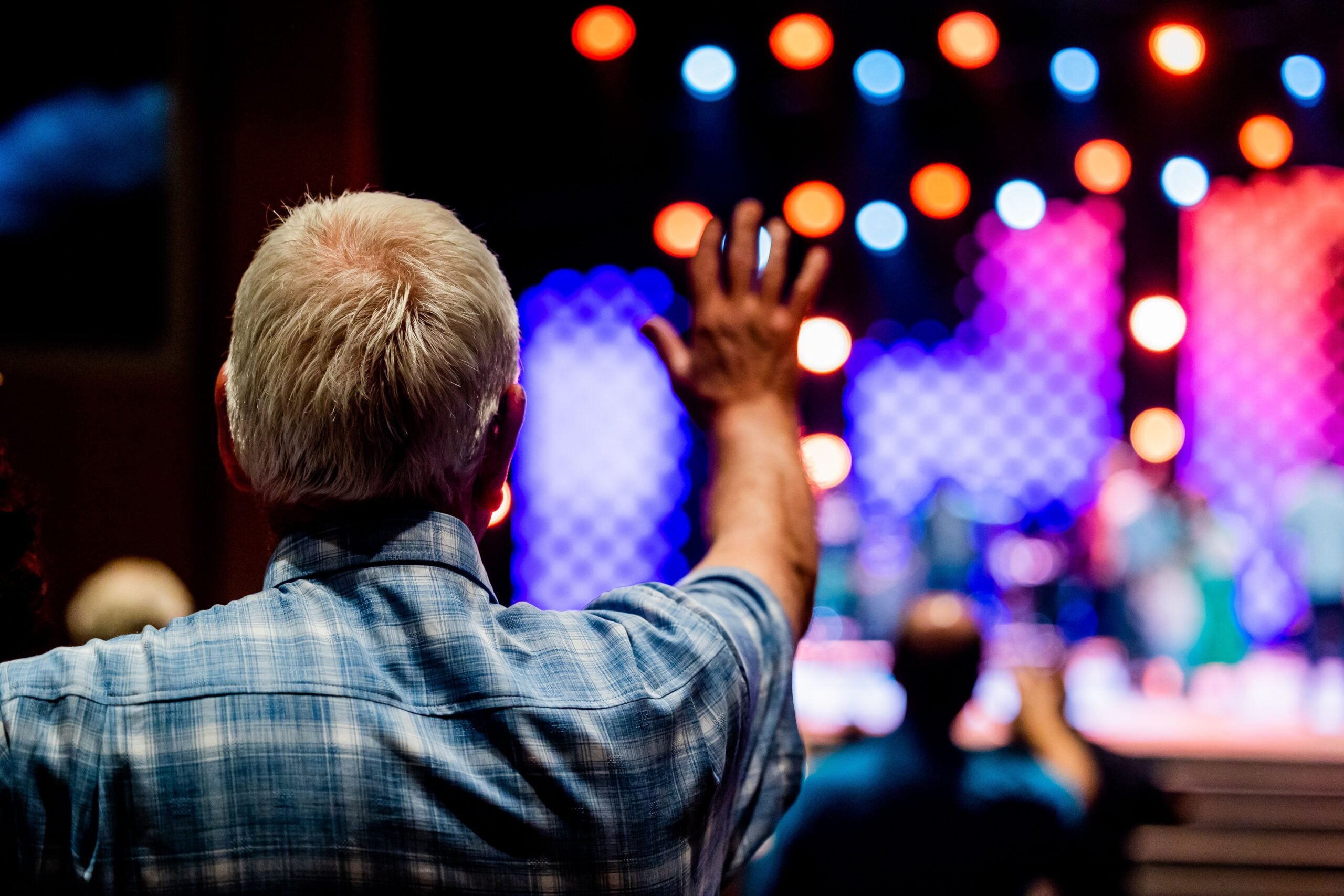 An older man with gray hair raises his hand in a crowd, perhaps eager to learn more about Boomers saving money, as he faces a brightly lit stage with colorful lights and blurred people in the background.