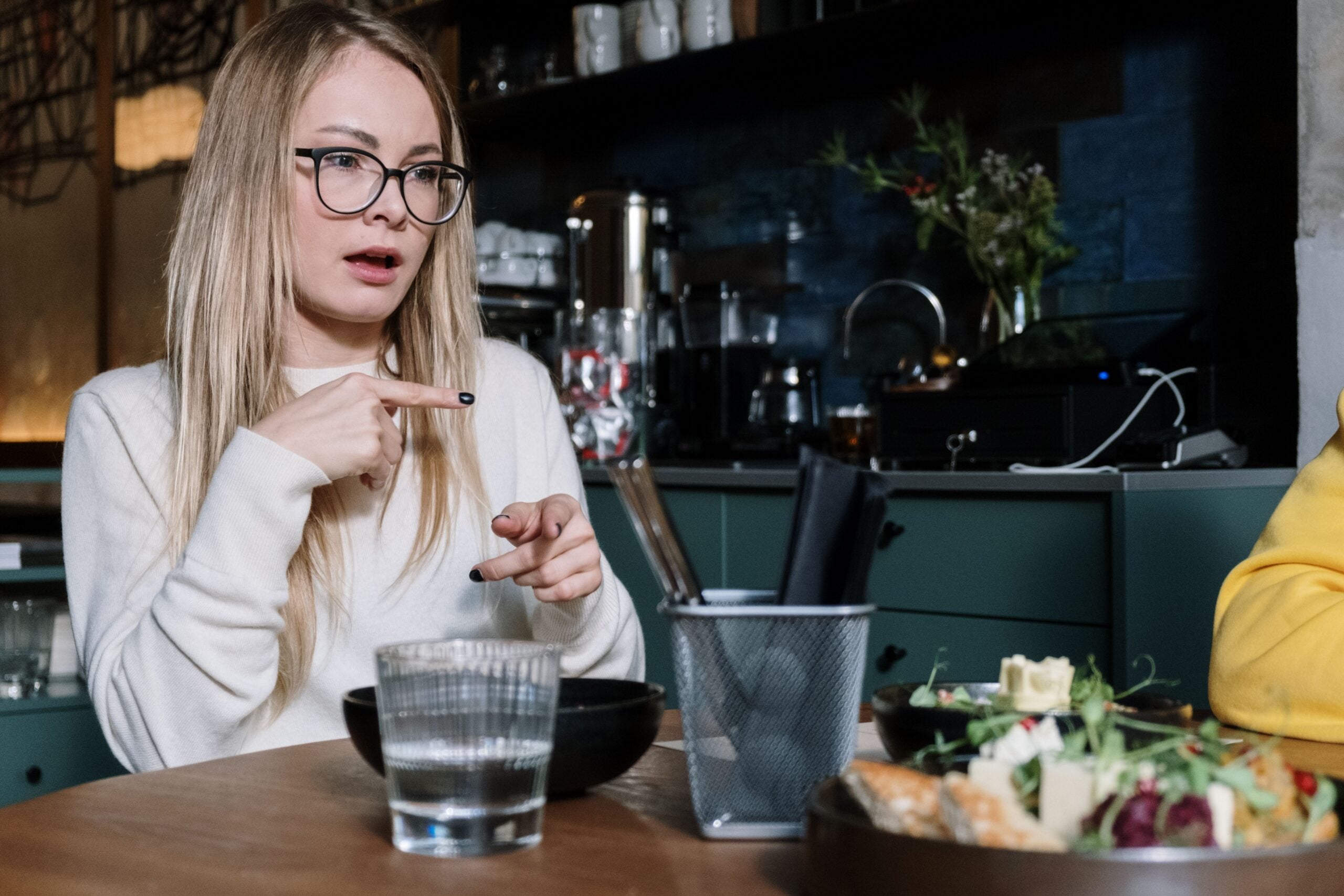 A woman with long blonde hair and glasses gestures while talking at a table set with food, a glass of water, and utensils in a modern kitchen or café setting.