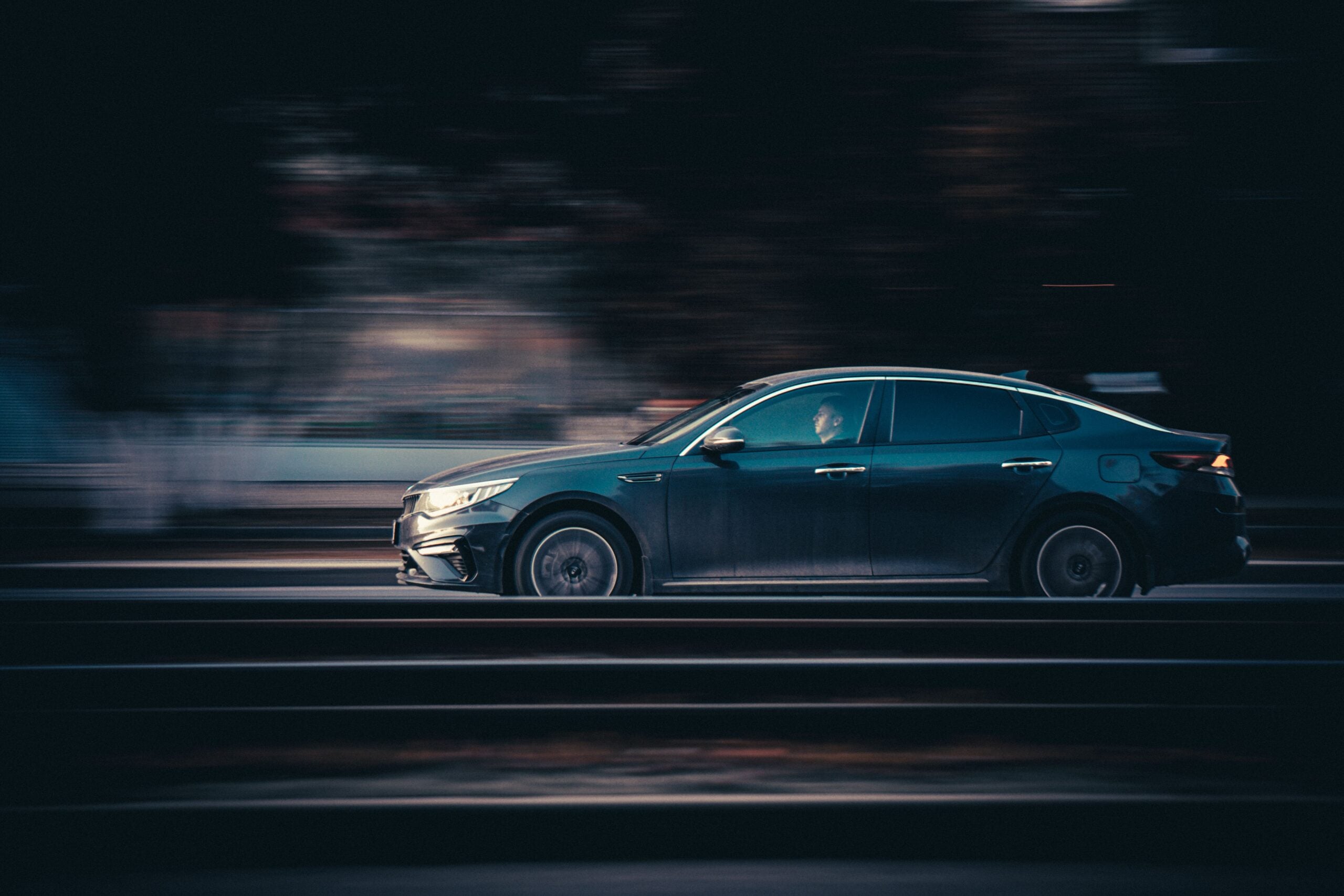 A dark-colored sedan is driving quickly on a road at dusk, with a blurred background indicating motion—perhaps reflecting the fast pace and modern challenges of life in 2026.