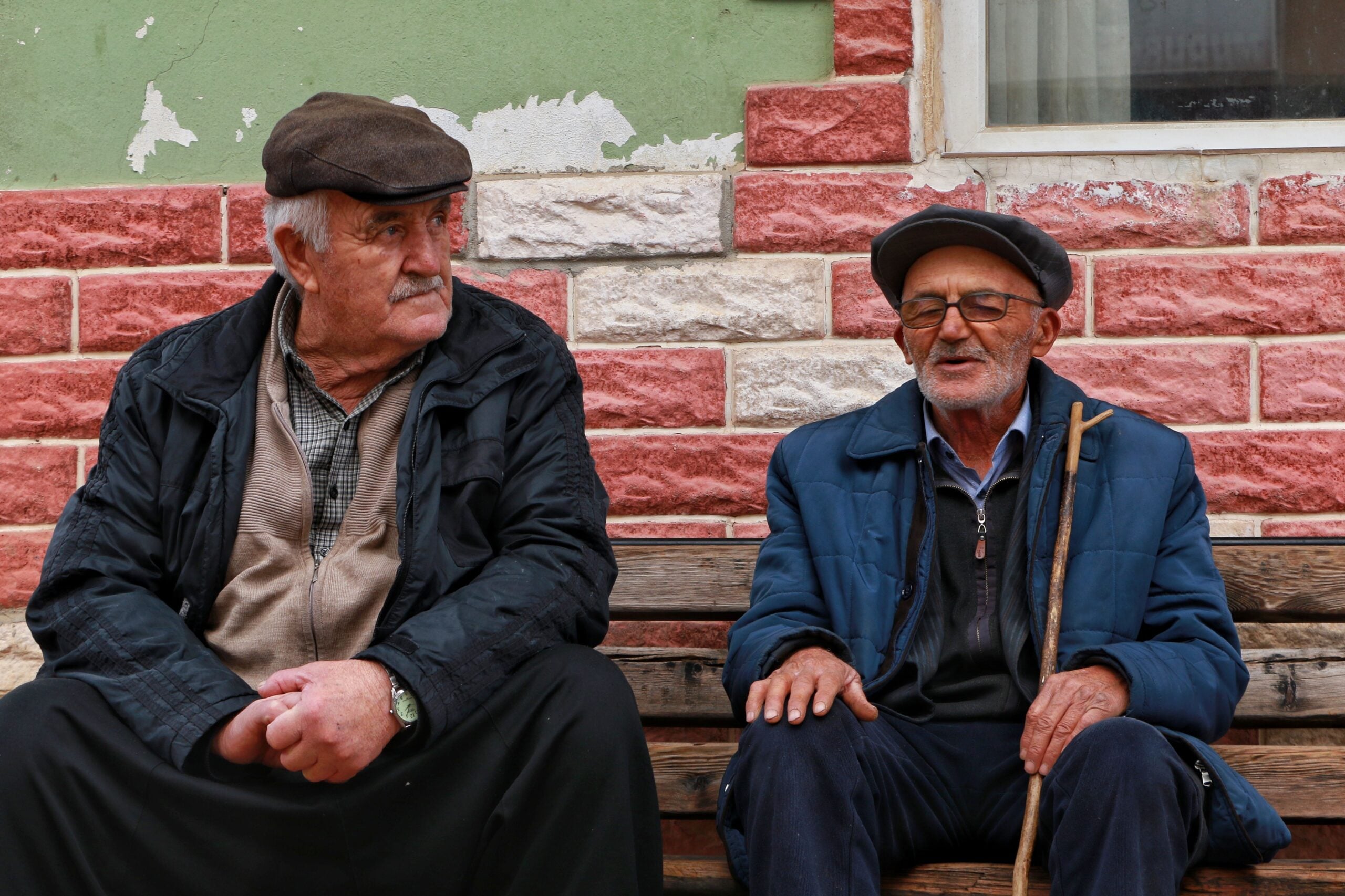Two elderly men sit on a wooden bench in front of a red and white brick wall. One man wears glasses and holds a cane, while both wear hats and jackets, appearing to be engaged in conversation—perhaps sharing some classic Boomer facts as they reminisce about days gone by.