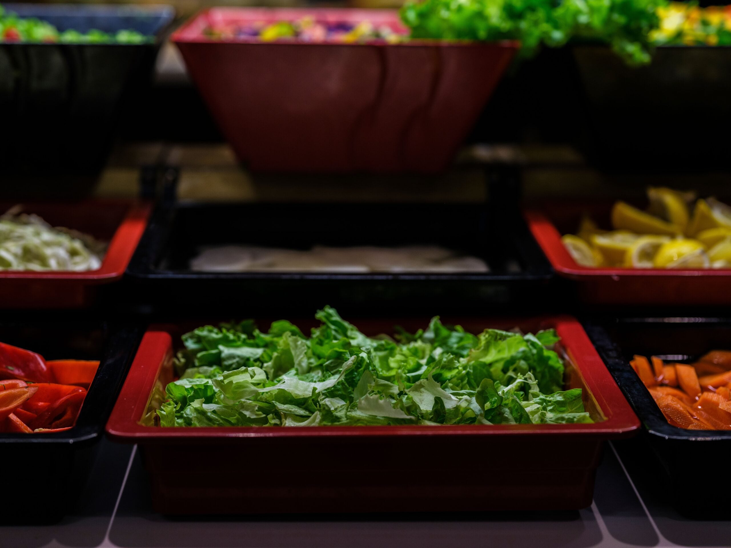 A close-up of a salad bar shows trays filled with fresh lettuce, sliced peppers, lemons, and other vegetables under bright lighting.