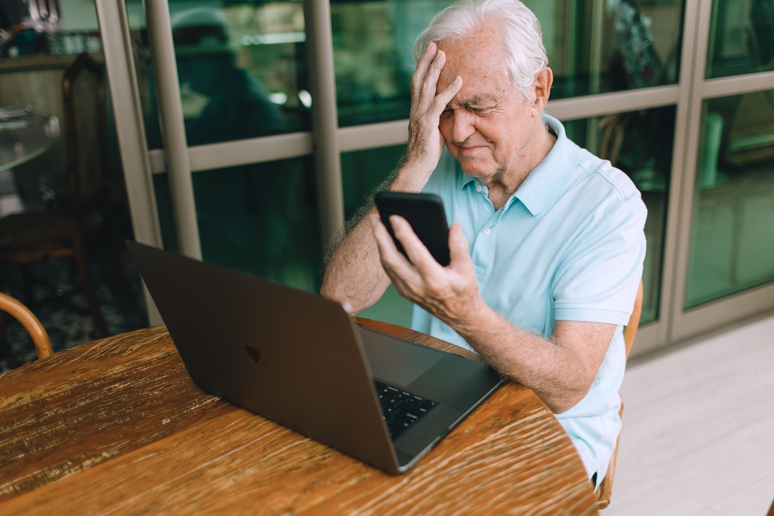 An elderly man sits at a table with a laptop, holding a smartphone and looking frustrated, with one hand on his forehead—struggling to navigate technology despite knowing all the boomer facts.