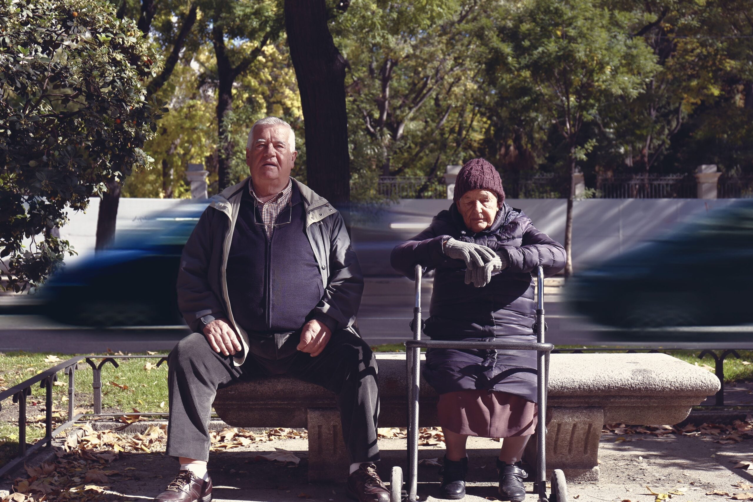 An elderly man and woman sit on a park bench, quietly enjoying the afternoon. The woman, using a walker and wearing a knit hat and gloves, shares Boomer facts with her companion as they watch trees sway and blurred cars pass by in the background.